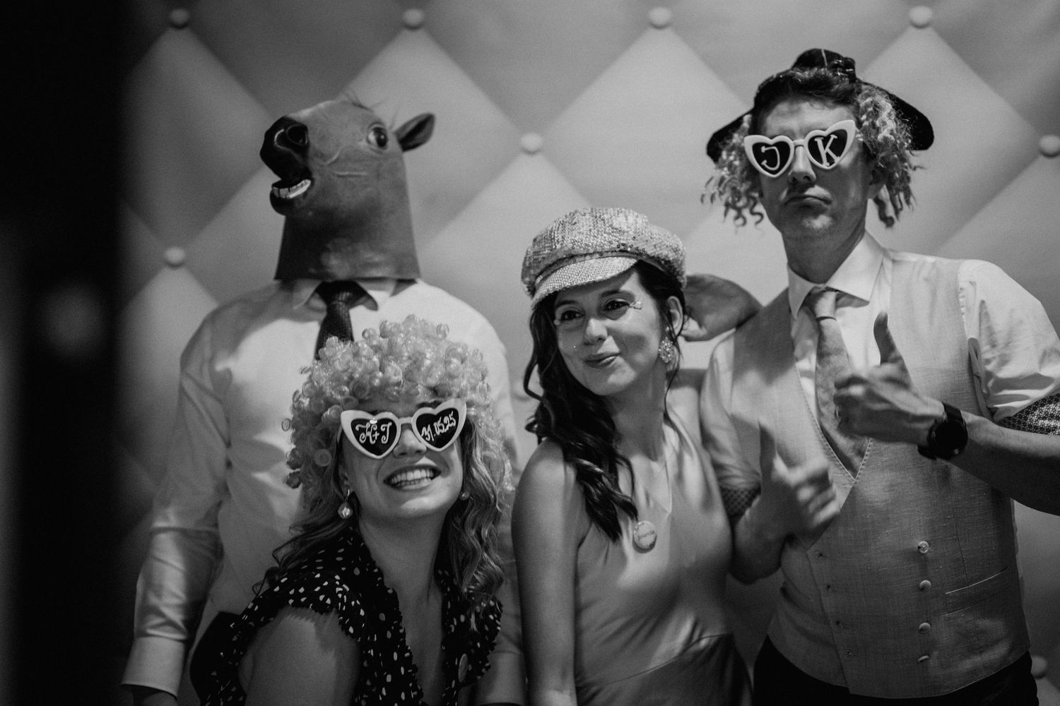 Black and white photo of four friends posing in a photo booth wearing fun props including a cow mask and novelty glasses.