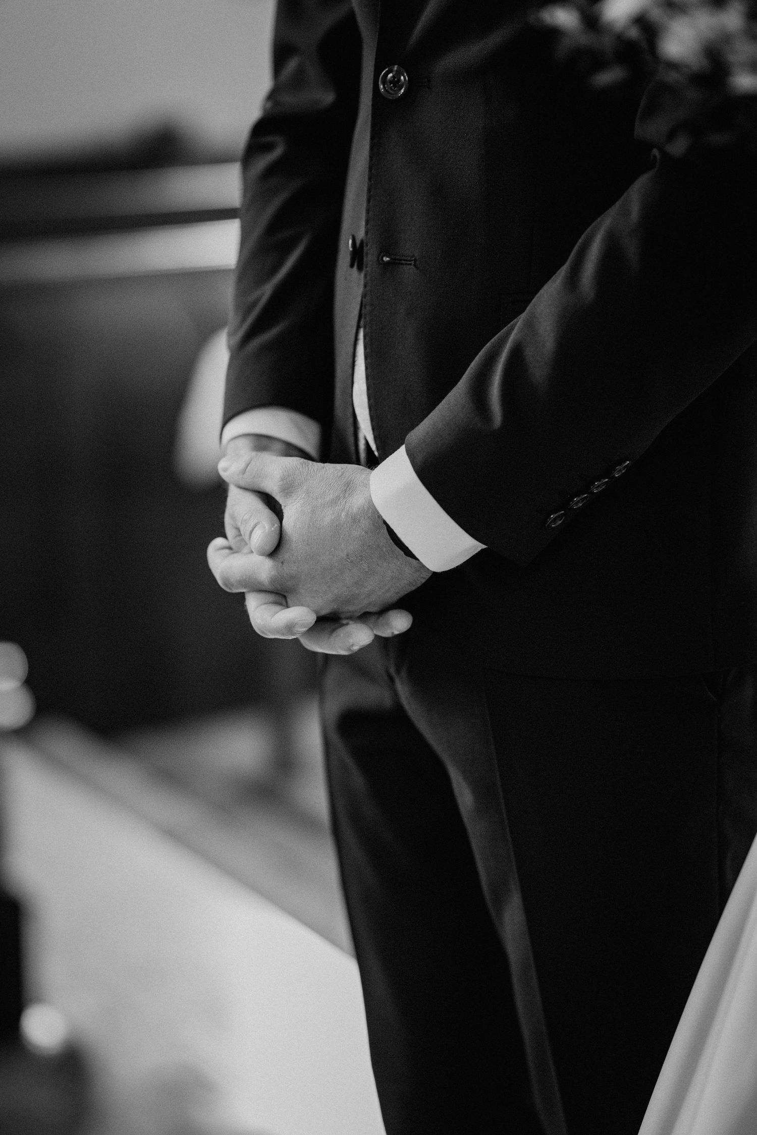 Black and white photo of a groom in a dark suit clasping his hands together during a wedding ceremony.