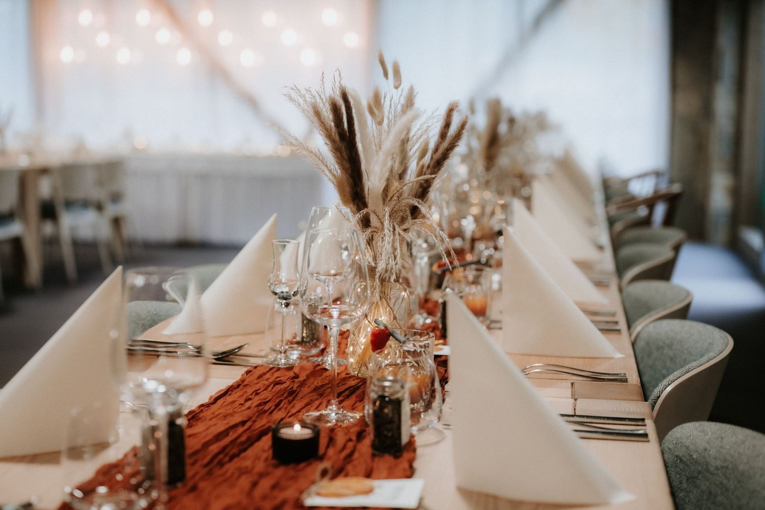 Elegant wedding reception table with pampas grass centerpieces, white napkins, and a rustic terracotta runner with candles.