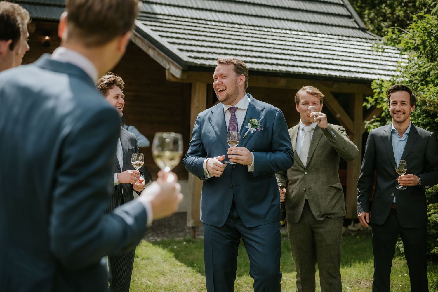 Groom laughing with groomsmen during outdoor wedding celebration, holding champagne glasses near rustic wooden building.