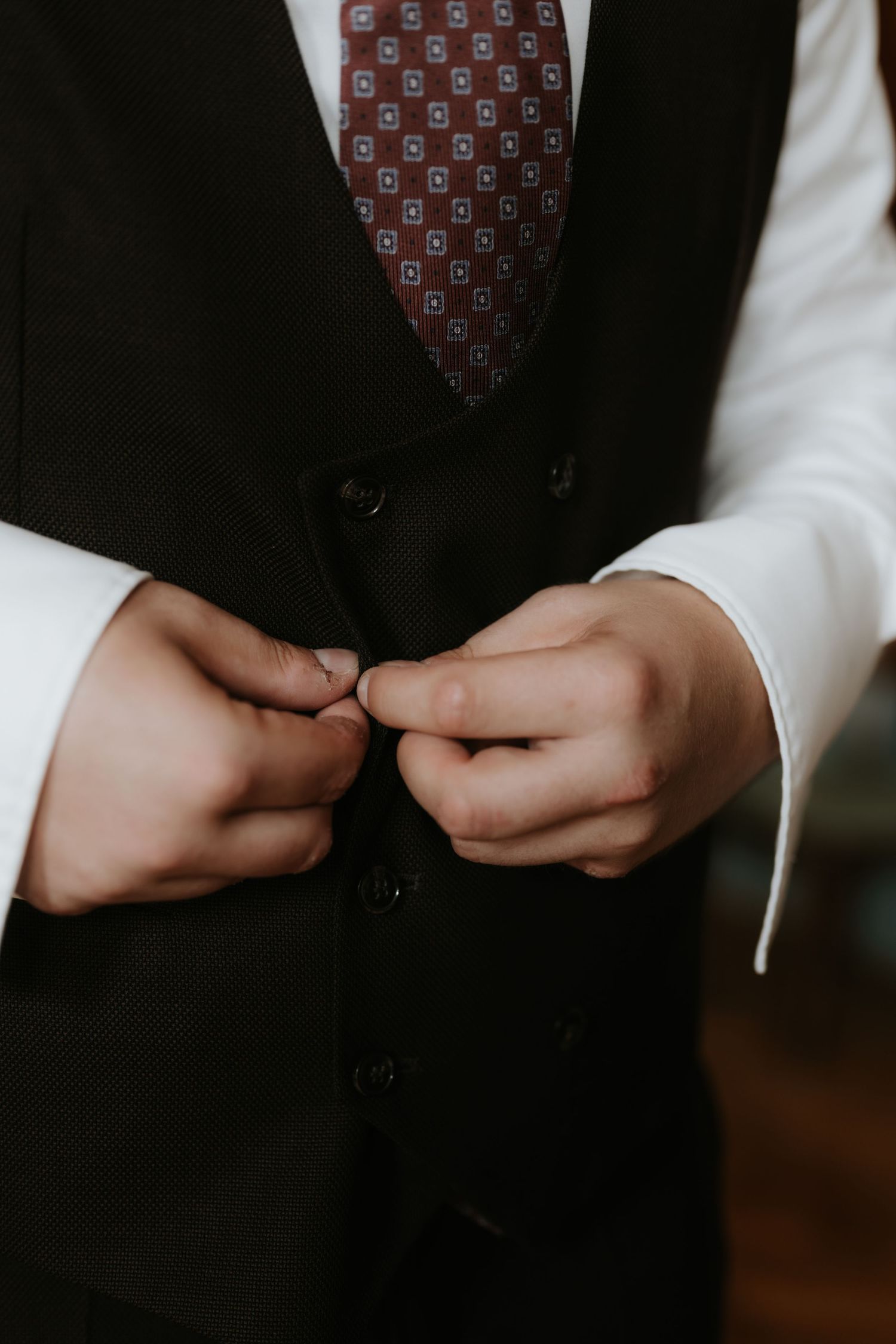 Man in black vest buttoning up over white shirt and burgundy patterned tie, formal wedding attire.