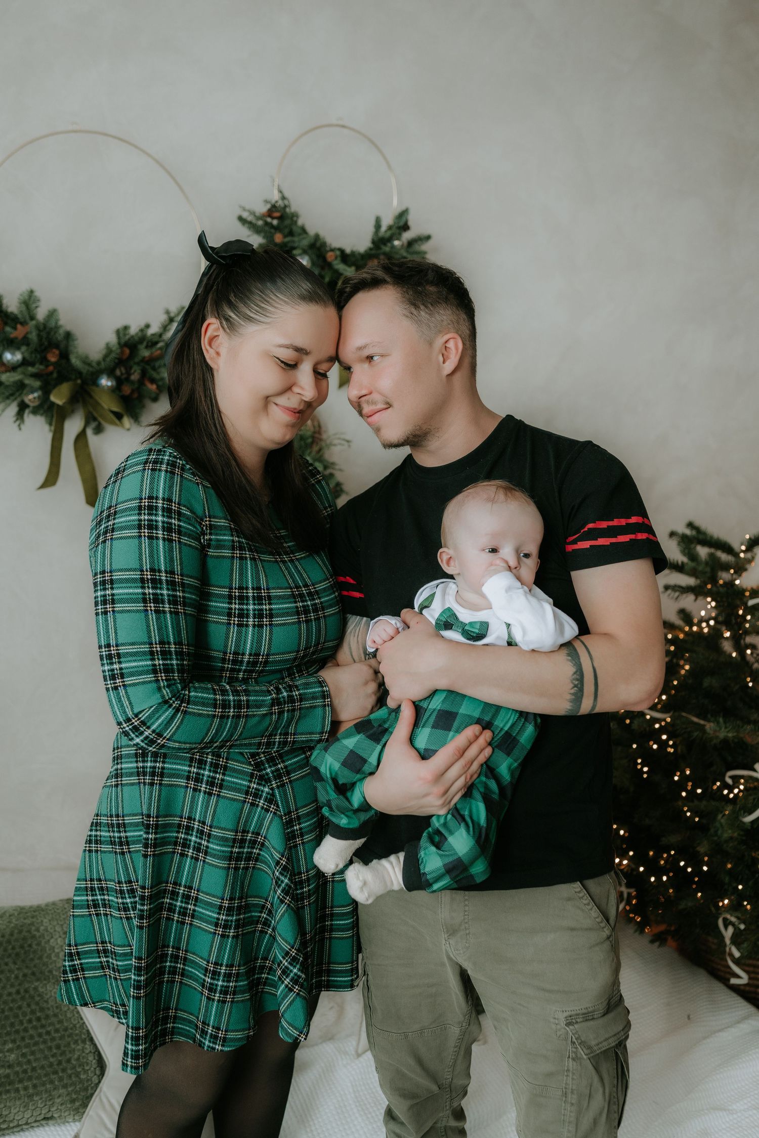 A young couple in matching green plaid outfits tenderly holds their newborn baby near Christmas tree decorations.