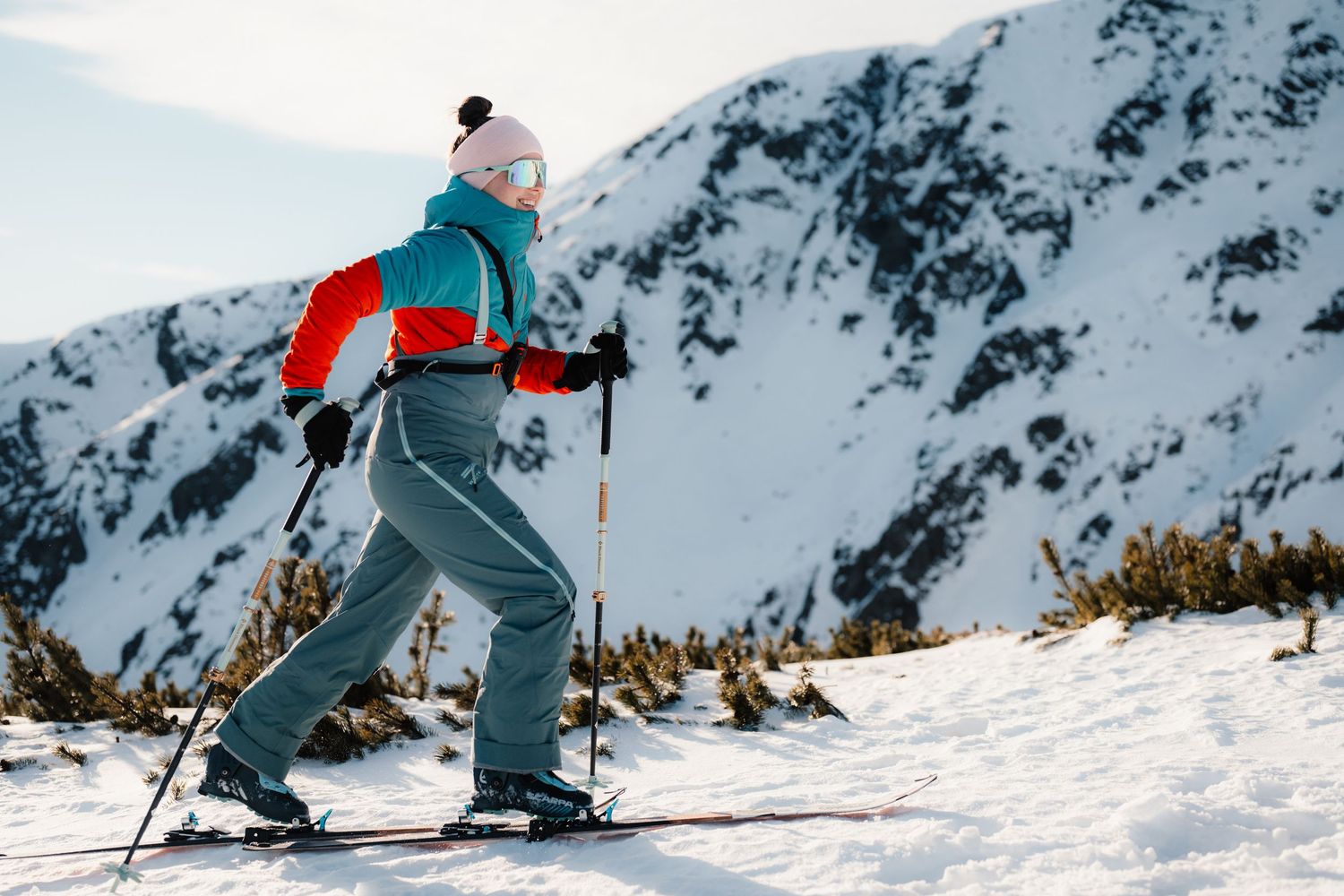 Woman cross-country skiing up a snowy mountain slope wearing colorful winter gear and goggles on a sunny day.
