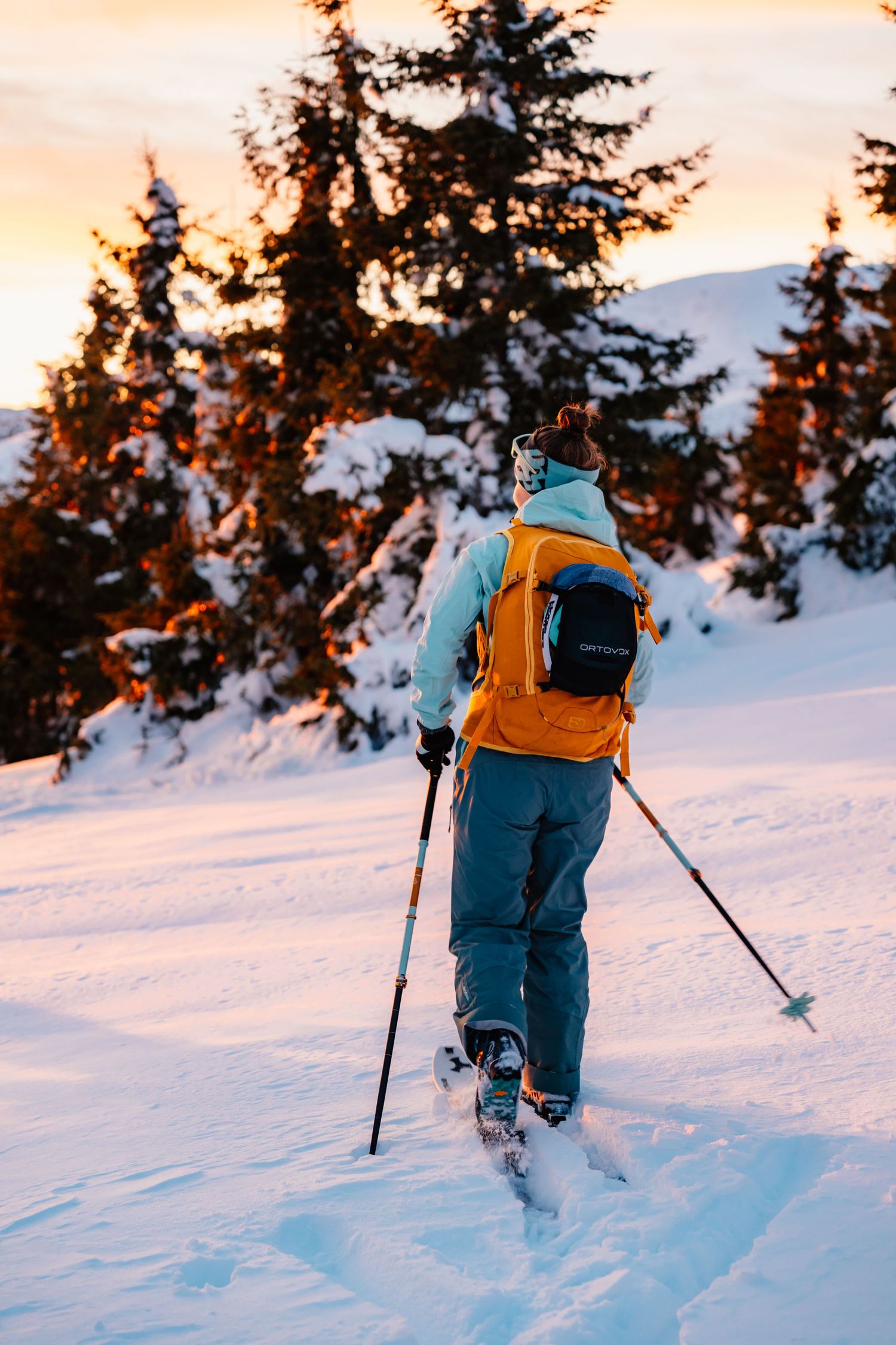 Back view of a snowshoer with orange backpack trekking through snowy forest at sunset.