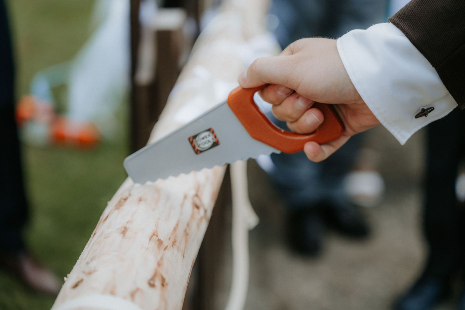 Person in formal attire sawing a wooden log with an orange-handled handsaw outdoors.