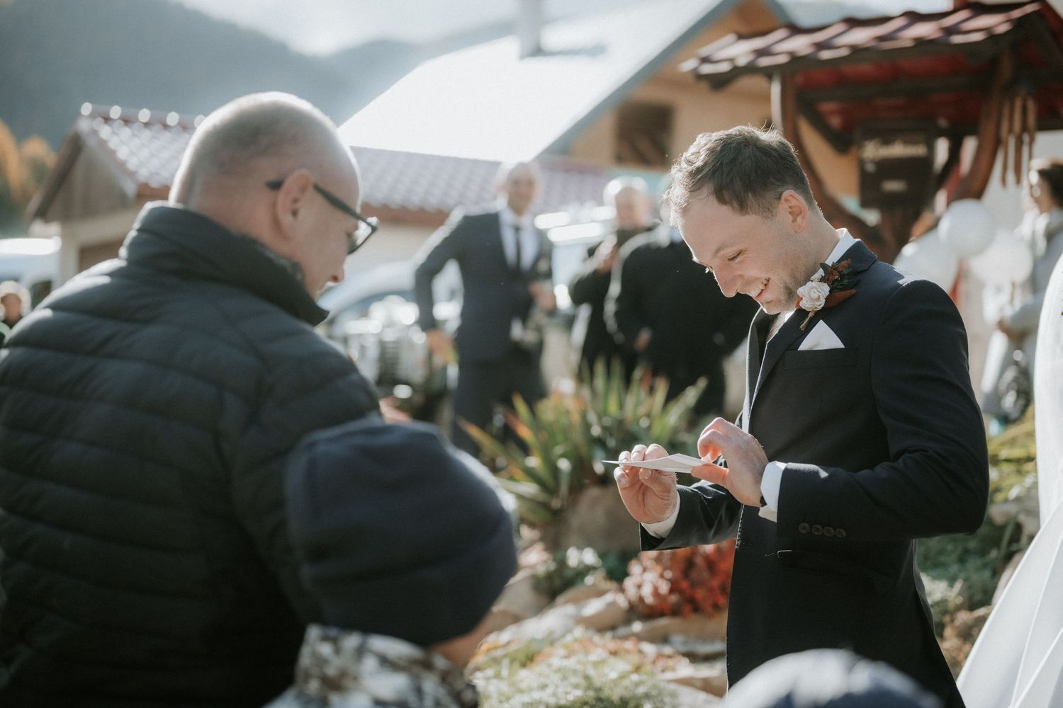 Groom in black tuxedo examining wedding ring outdoors at winter wedding ceremony with guests nearby.