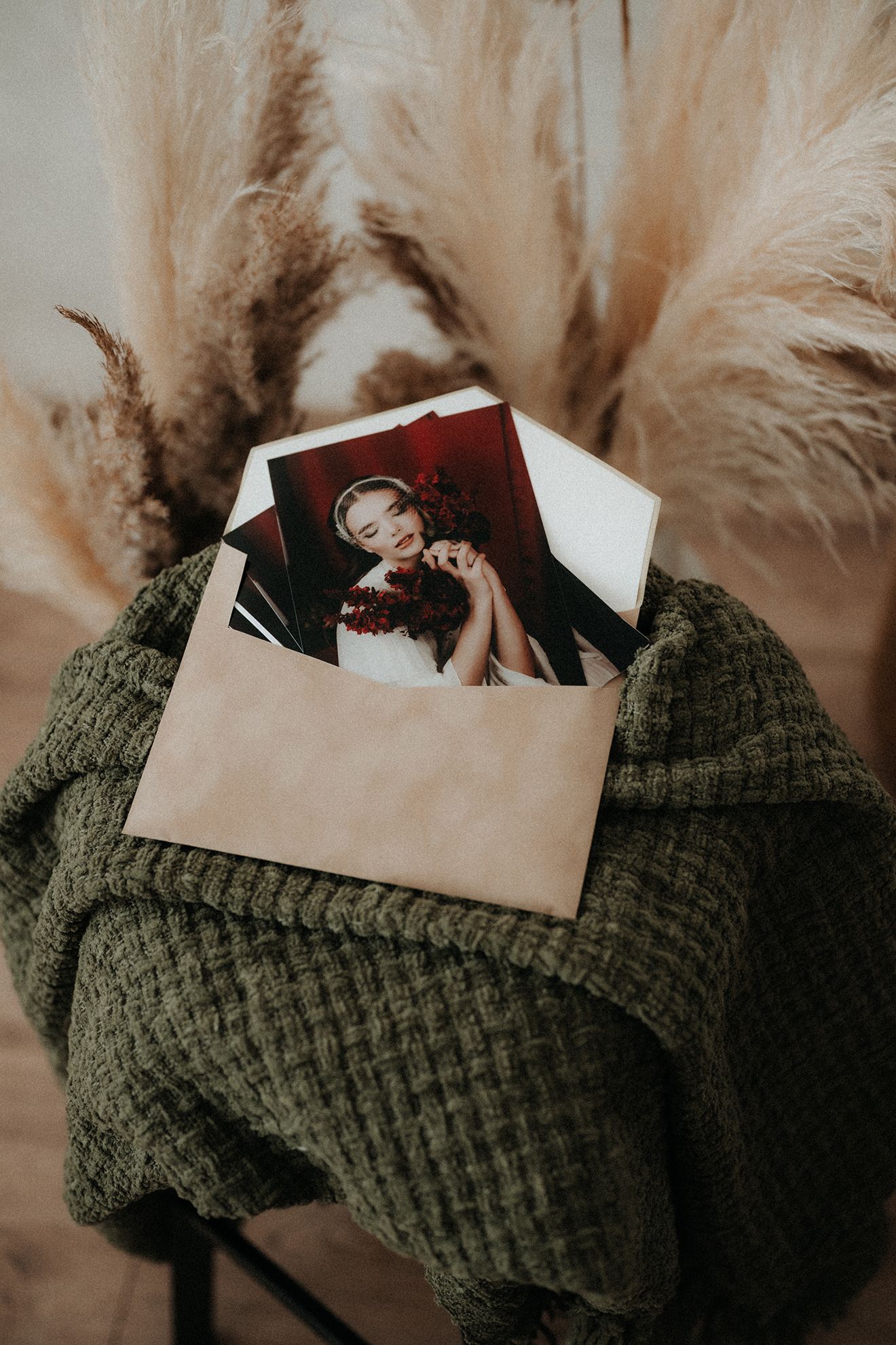 Polaroid photos in a kraft envelope wrapped in a green knit blanket with dried pampas grass in the background.