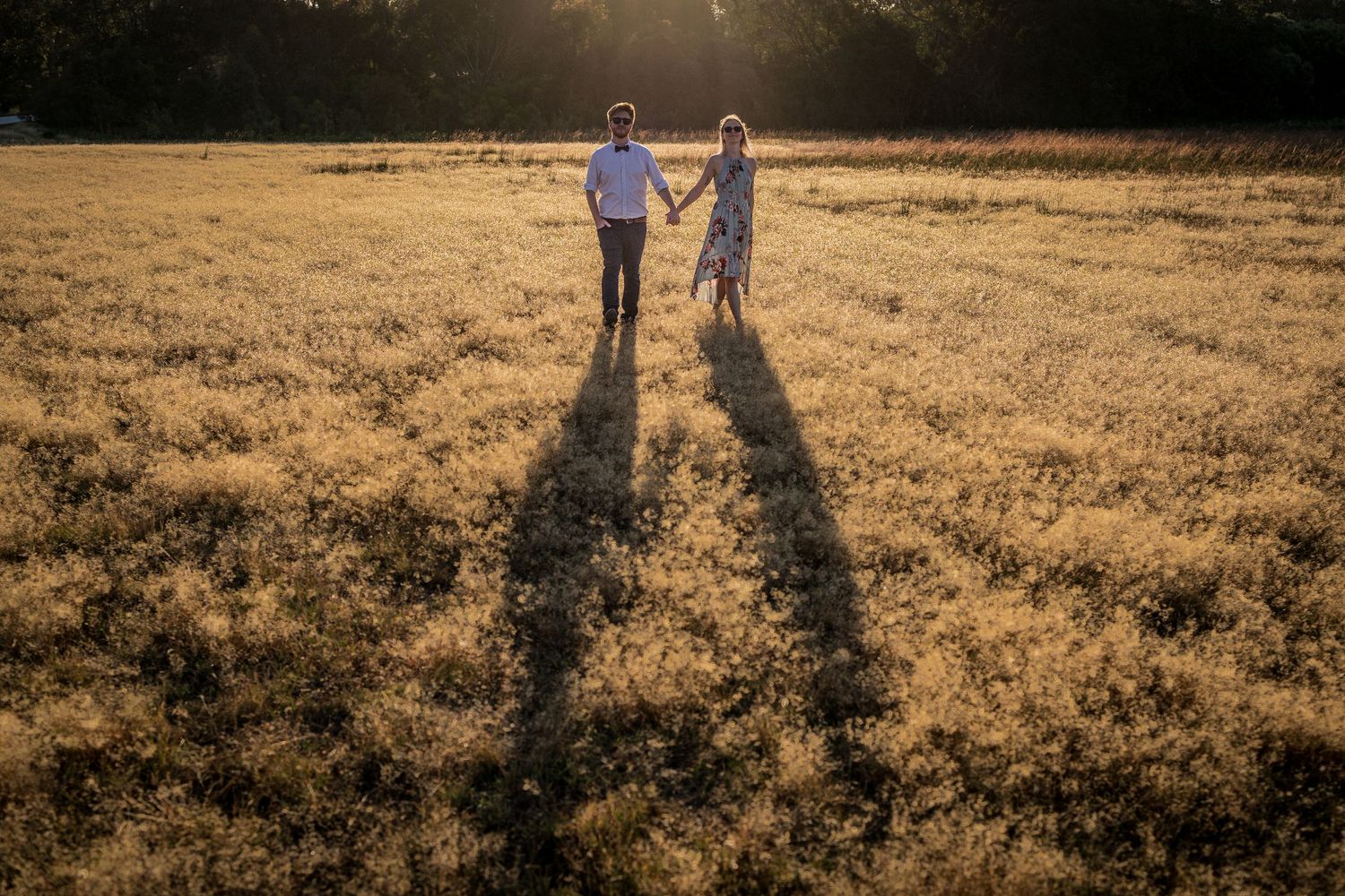 Couple holding hands in golden field at sunset with long shadows stretching across grass.