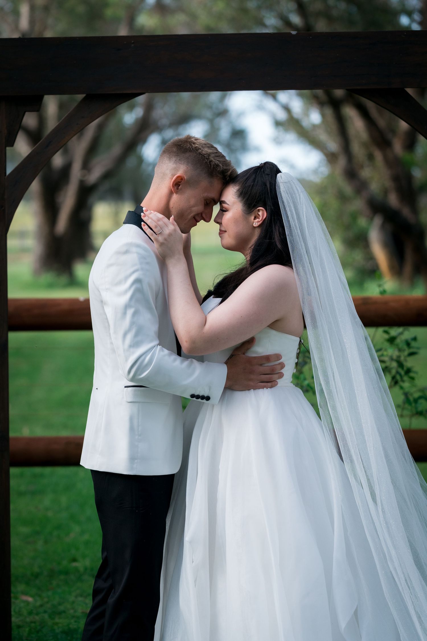 A bride and groom share an intimate moment under a wooden arch surrounded by lush green trees.