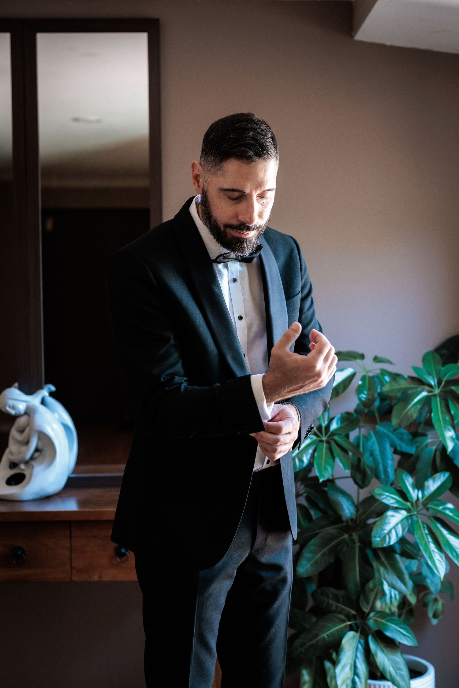 A well-dressed groom in a black tuxedo with bow tie adjusts his cufflinks while preparing for his wedding day.