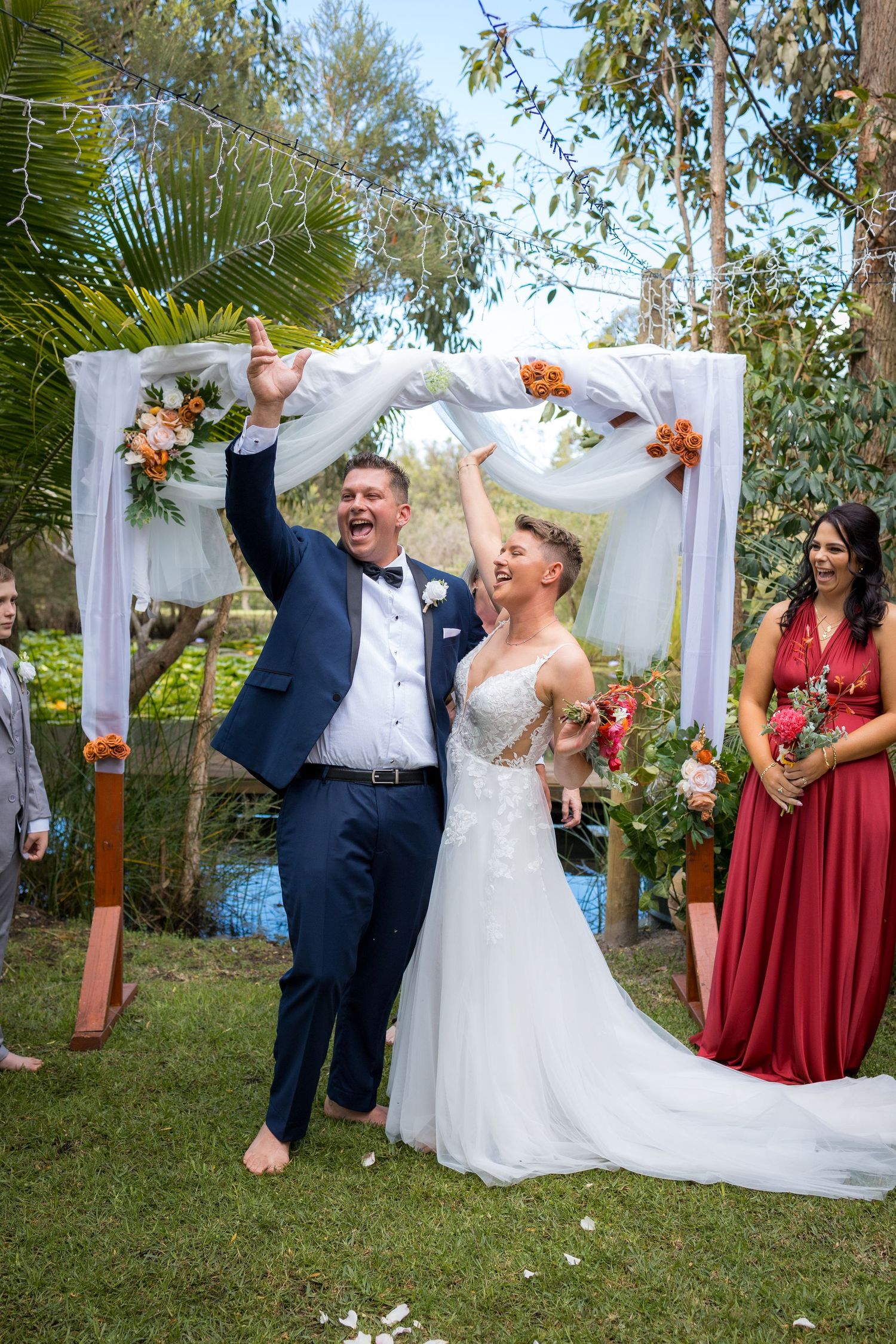 Wedding party celebrates joyfully under a white draped arch decorated with orange and peach florals in an outdoor garden setting.