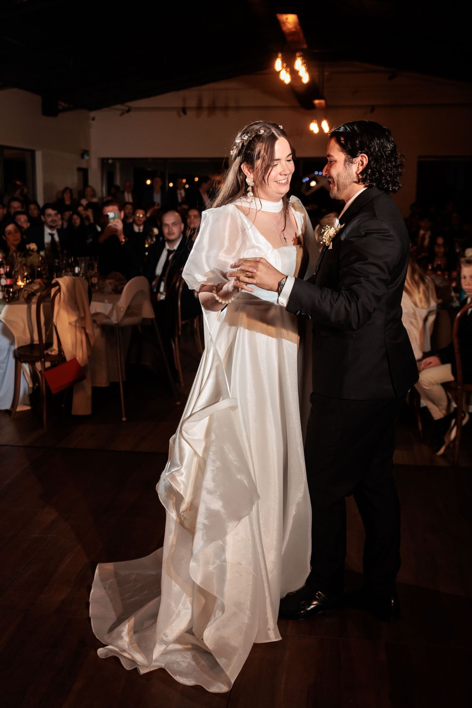 Bride and groom sharing first dance, bride laughing in white satin gown at wedding reception.