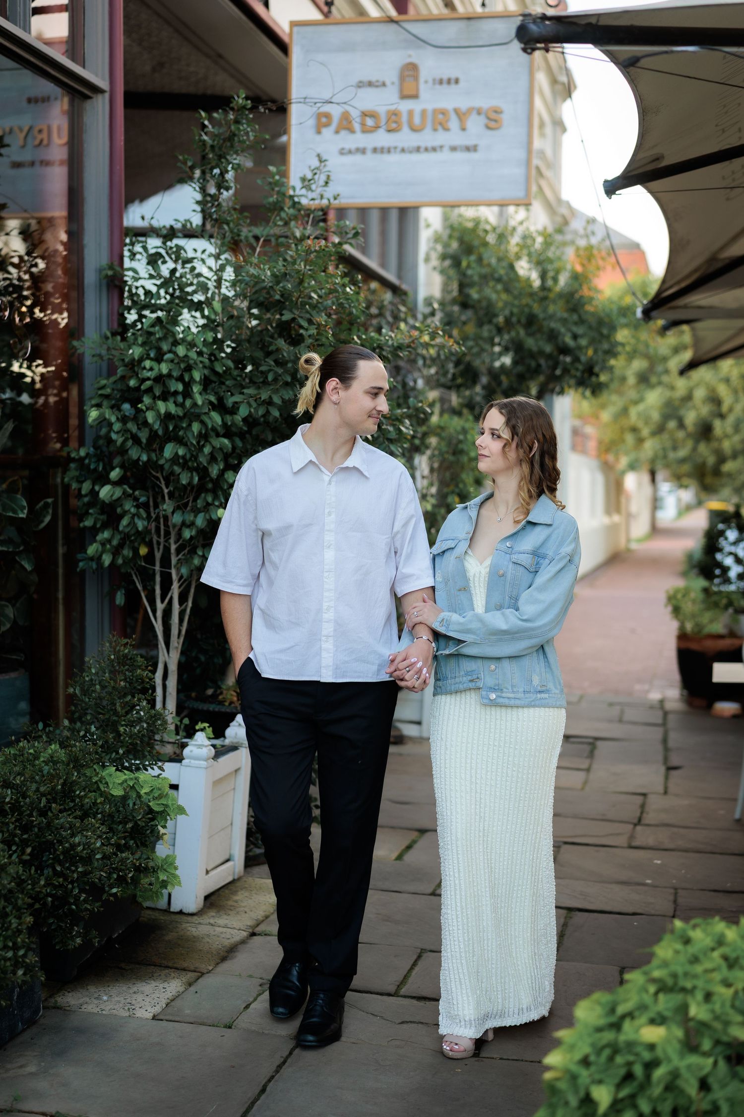 Couple holding hands and smiling outside Padbury's restaurant, man in white shirt, woman in denim jacket and maxi dress. at Guildford, Perth
