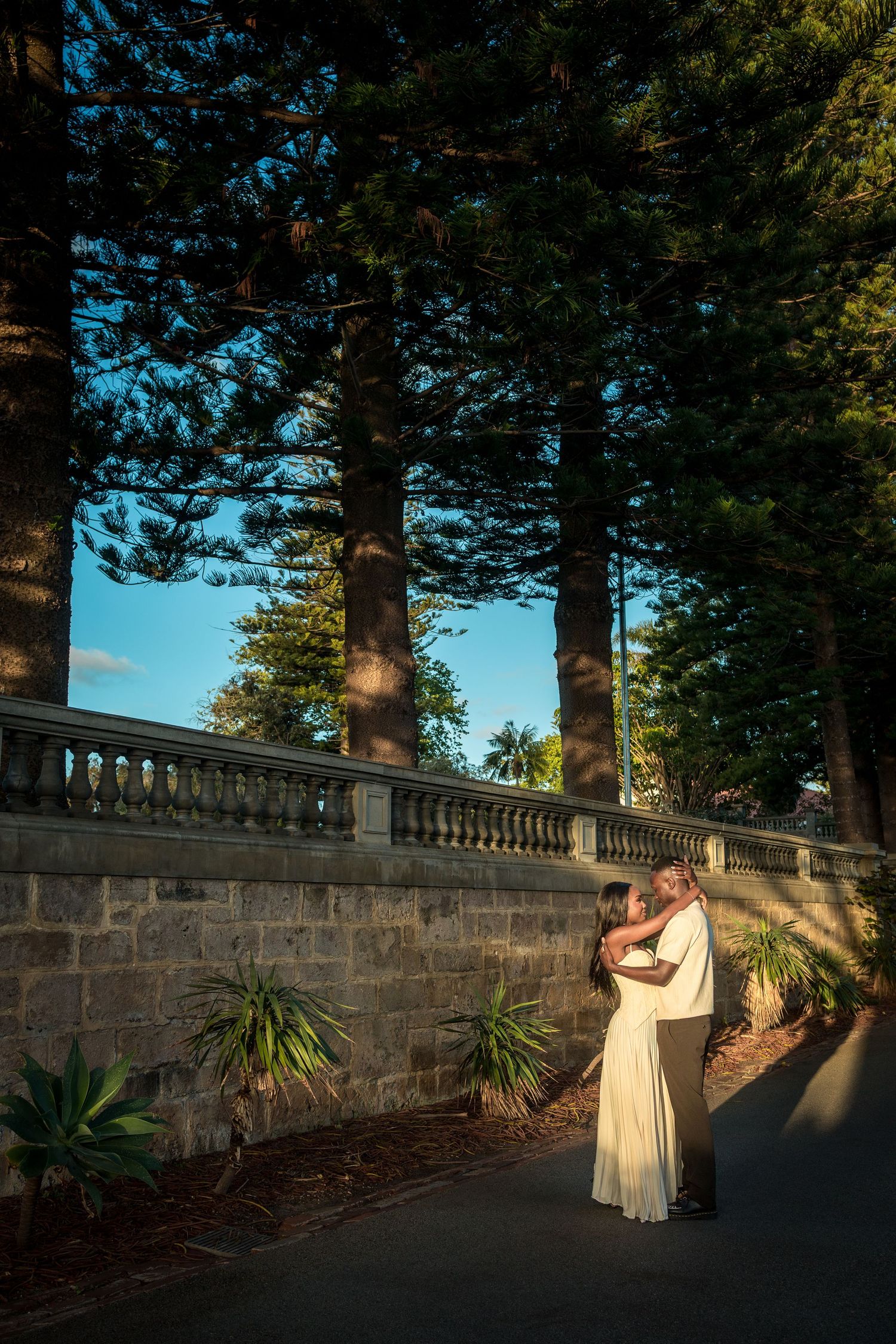 Couple embracing at golden hour beside a historic stone balustrade wall, with tall Norfolk pines and agave plants under a blue sky.