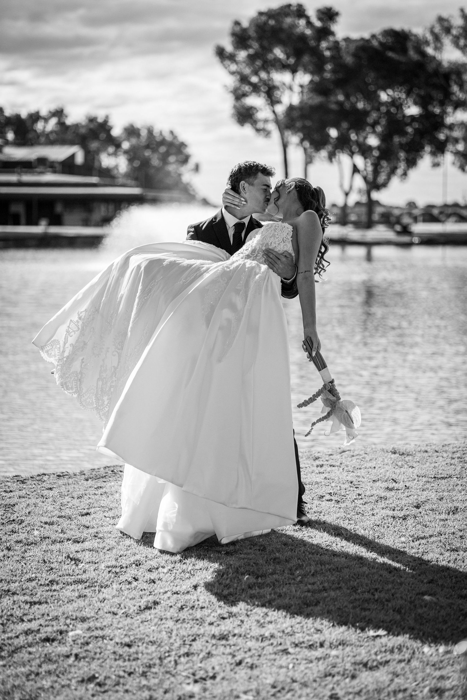Groom lifts bride in white gown for a romantic kiss outdoors by a lake in a classic black and white wedding photo.