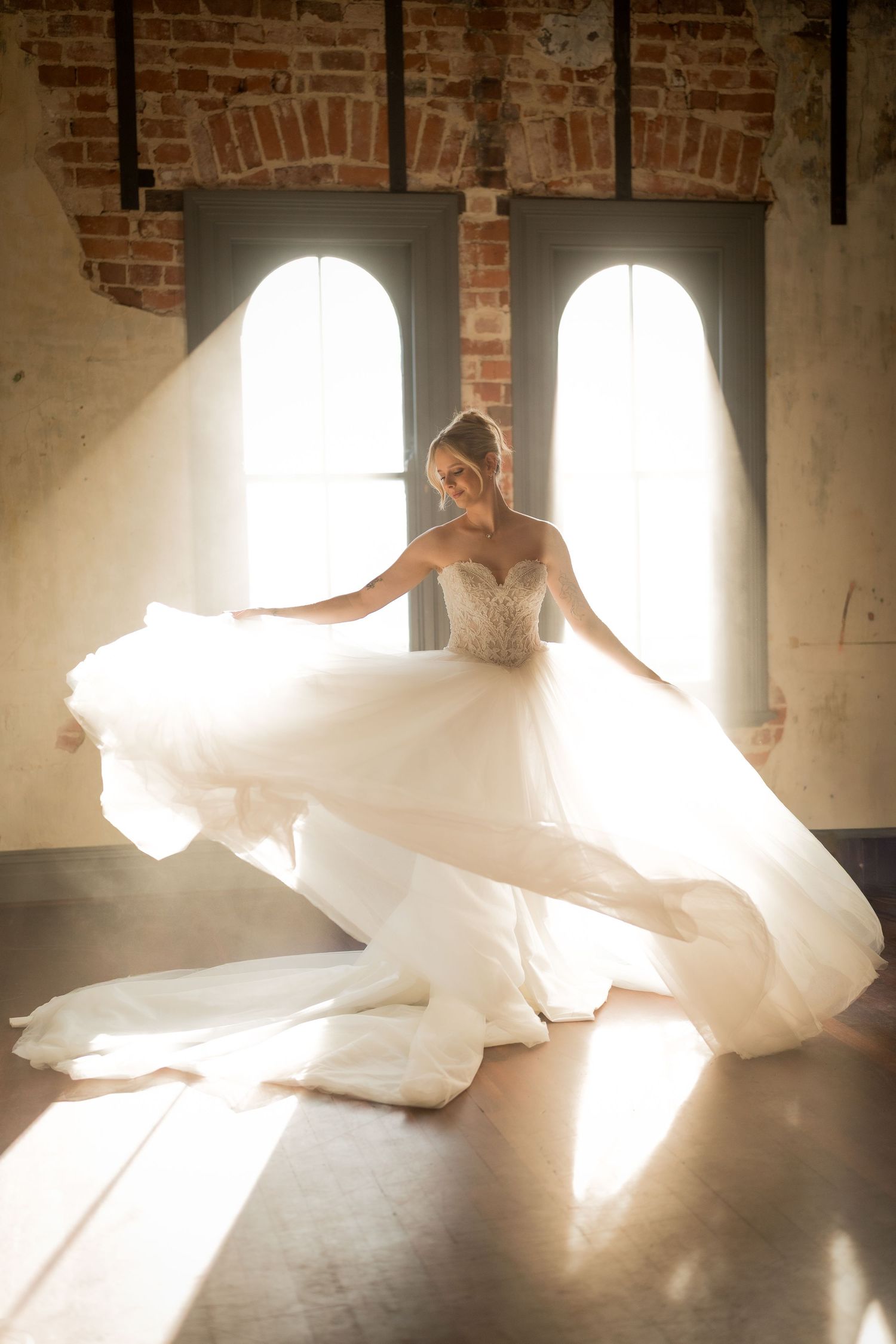 Bride twirling in a flowing white ball gown wedding dress, bathed in natural light from arched windows in a rustic brick venue.