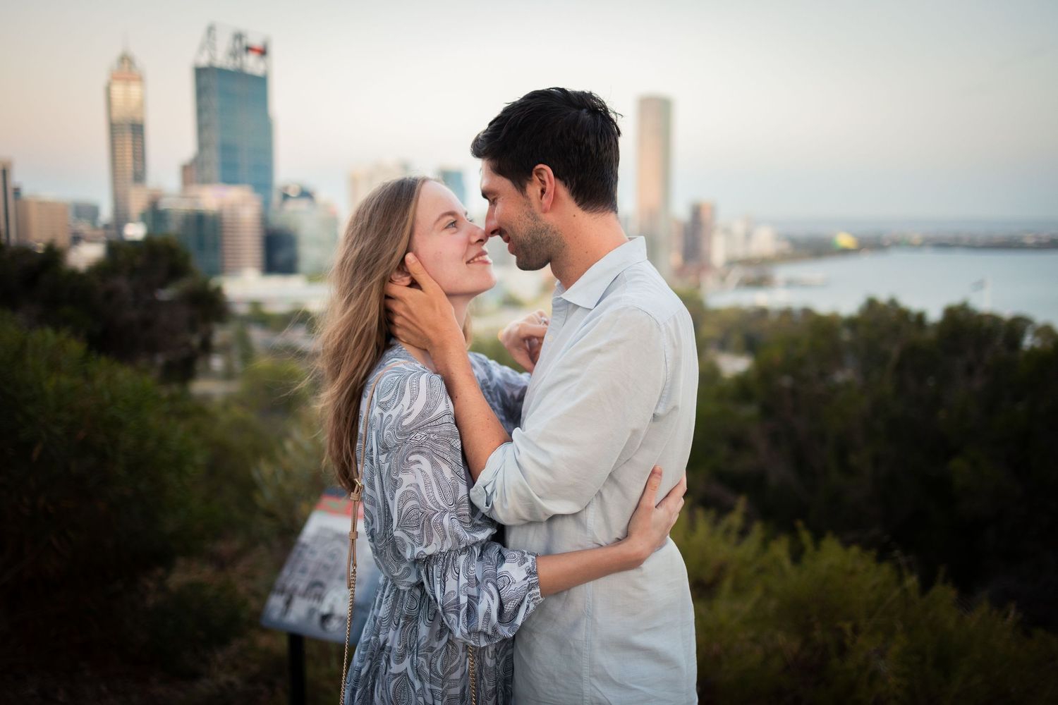 Romantic couple embracing with city skyline and river views in background at golden hour.