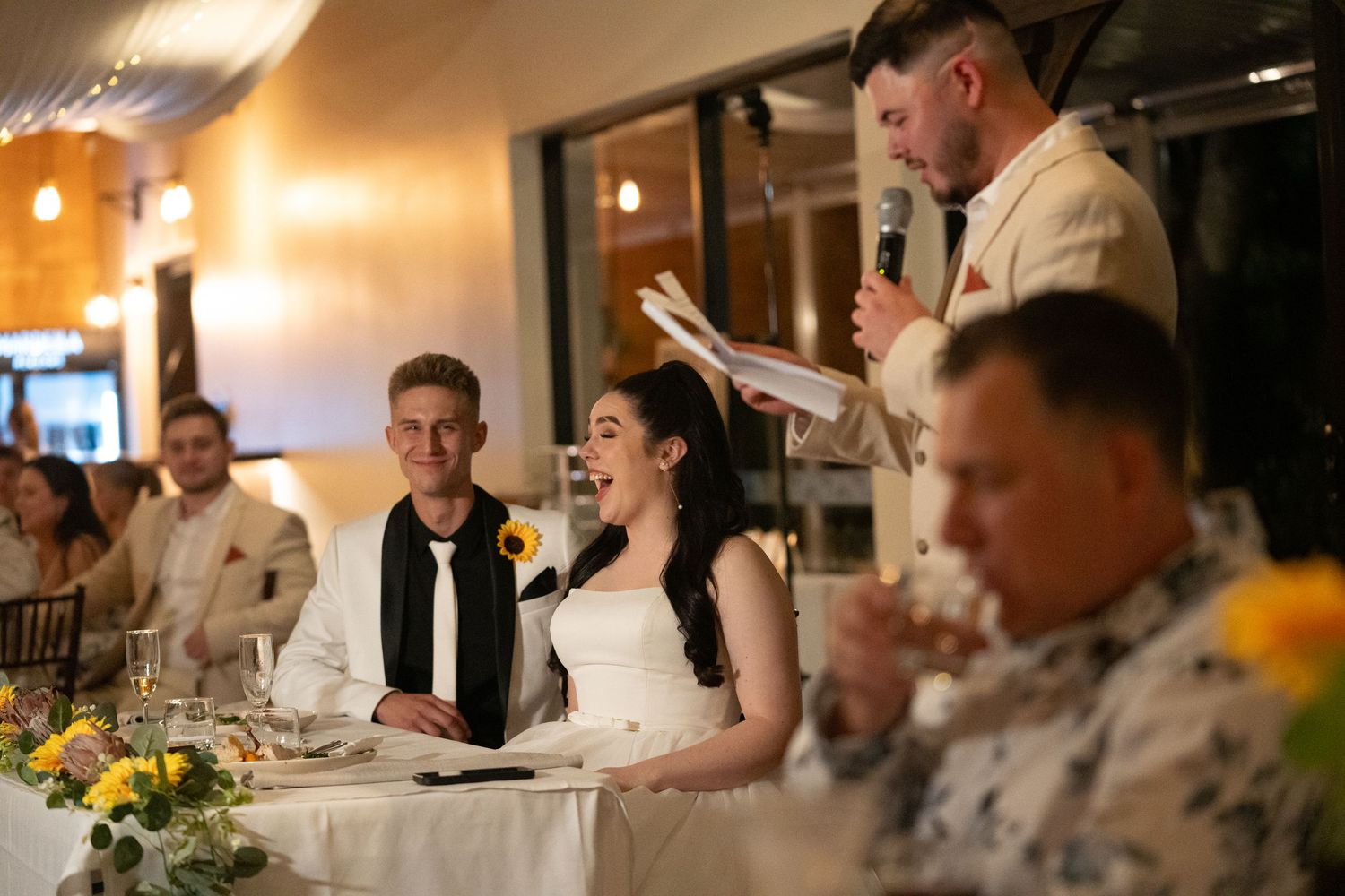Wedding reception dinner with guests seated at decorated tables in warm lighting.