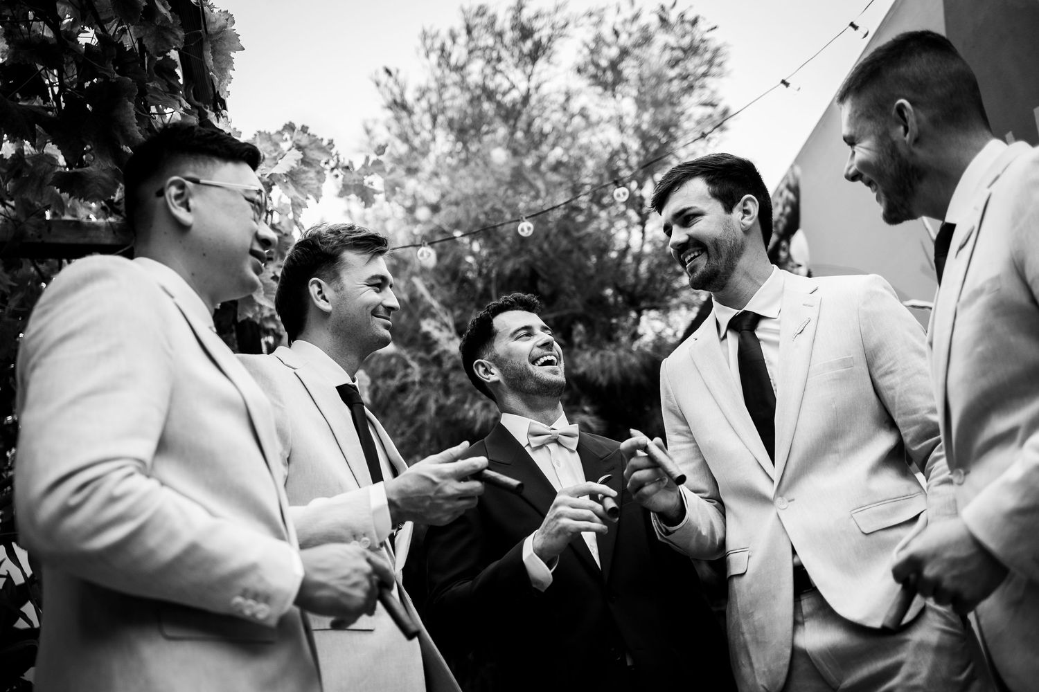 Five groomsmen in suits laughing together outdoors at a wedding celebration.