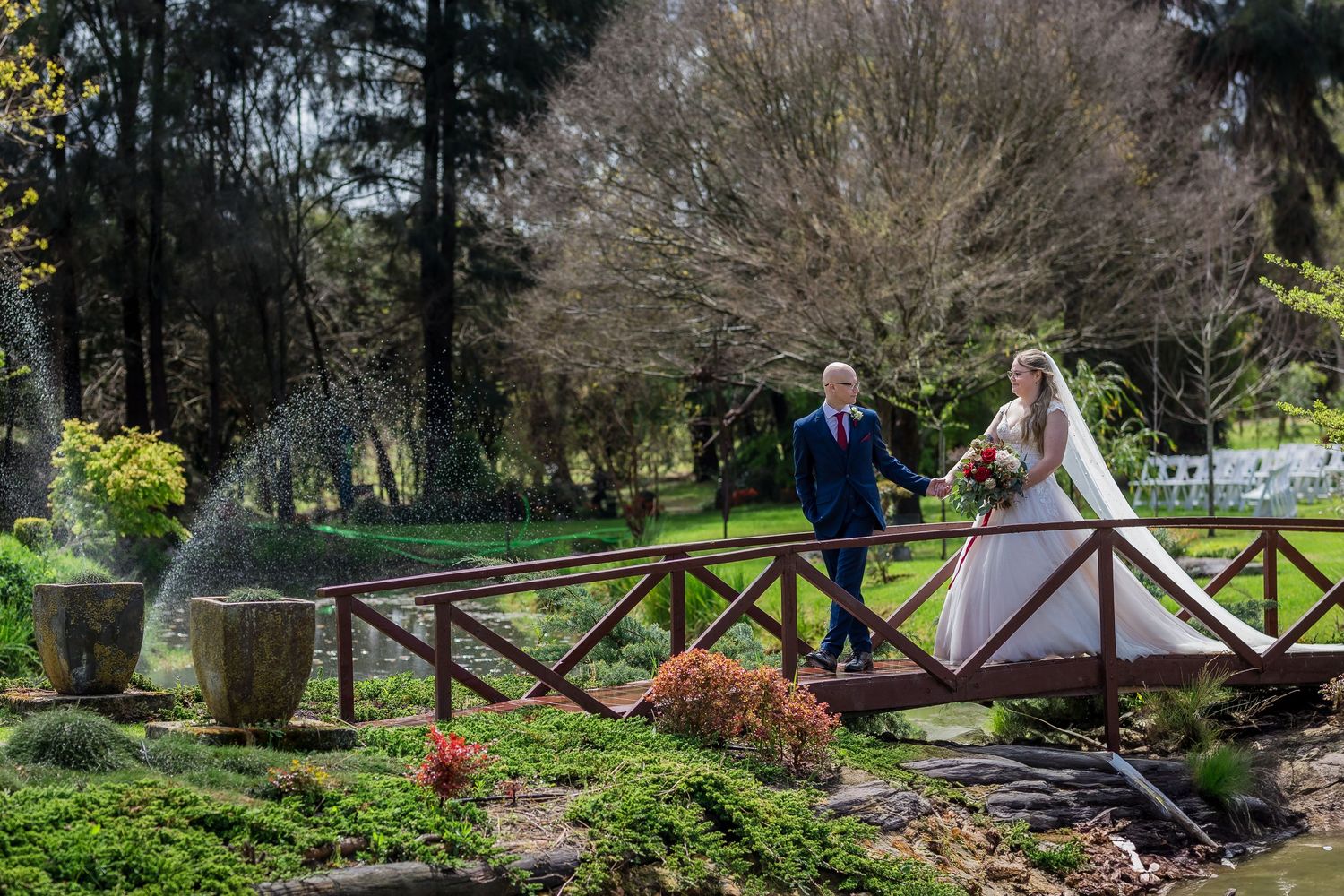 Bride and groom walking hand in hand on a wooden bridge surrounded by lush gardens with a fountain at Core Cider.
