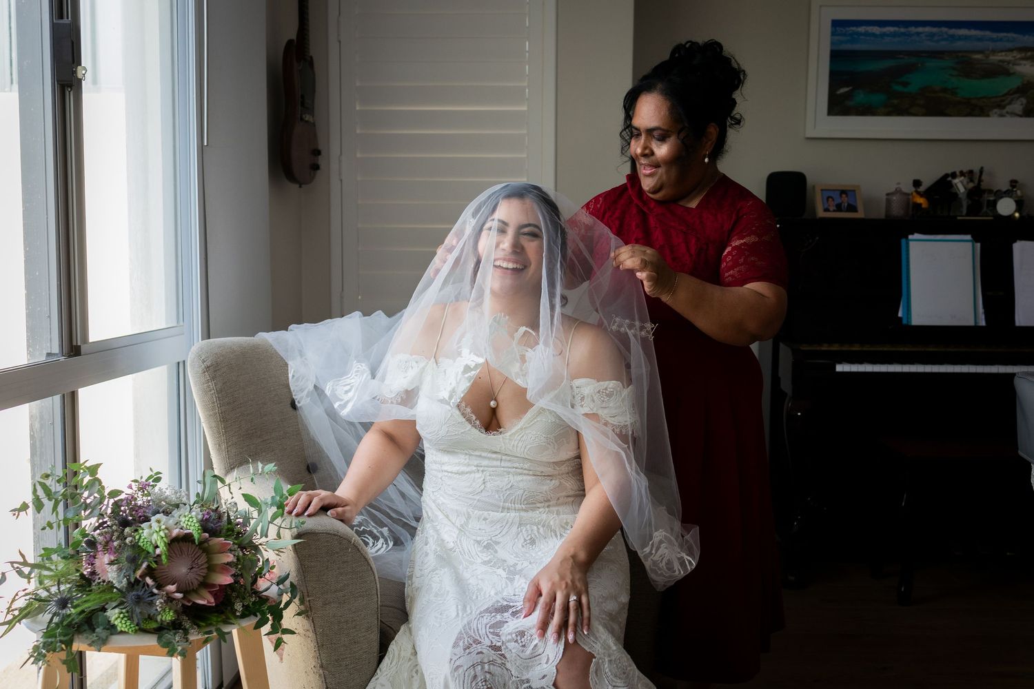 Bride in lace wedding dress and veil laughing as bridesmaid in red dress adjusts her veil before the ceremony.
