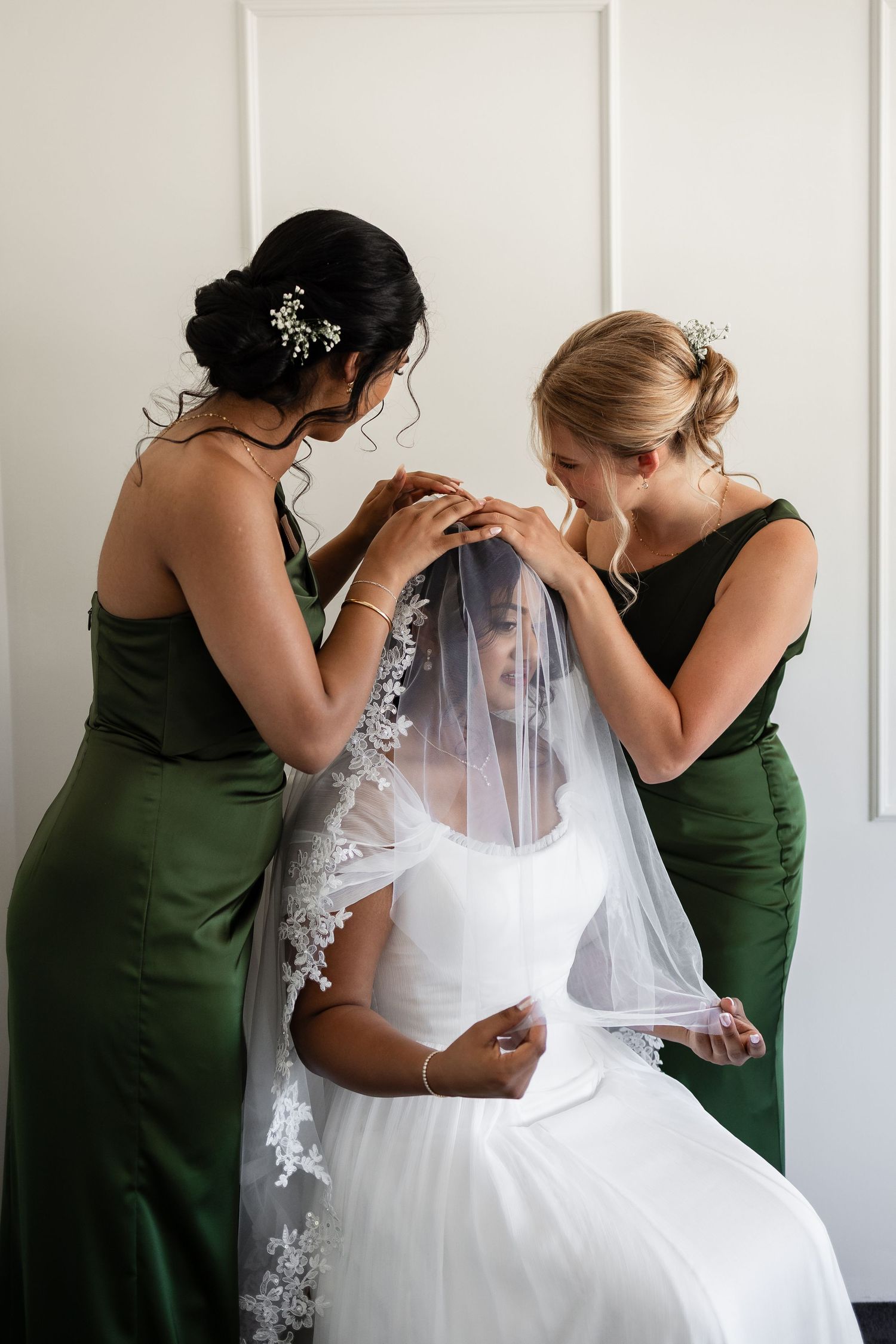 Two bridesmaids in green satin dresses carefully arrange lace-trimmed veil on seated bride in white wedding gown.