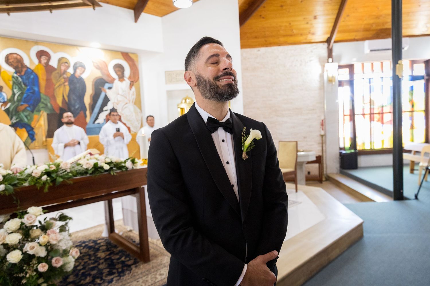 Emotional groom in black tuxedo reacts as bride walks down the aisle in a decorated church ceremony.