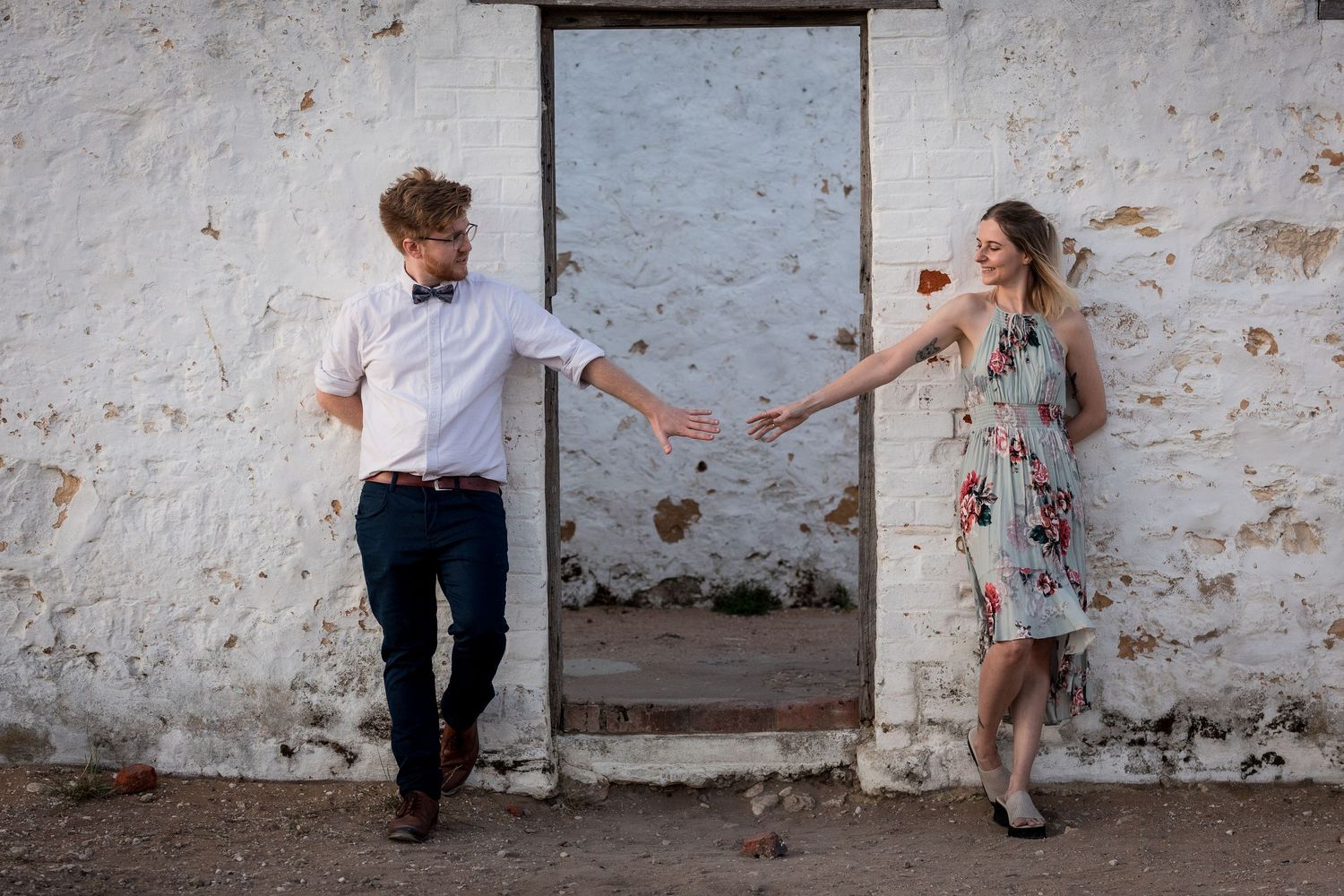 Couple reaching toward each other in front of a rustic white weathered wall, dressed in elegant casual attire.