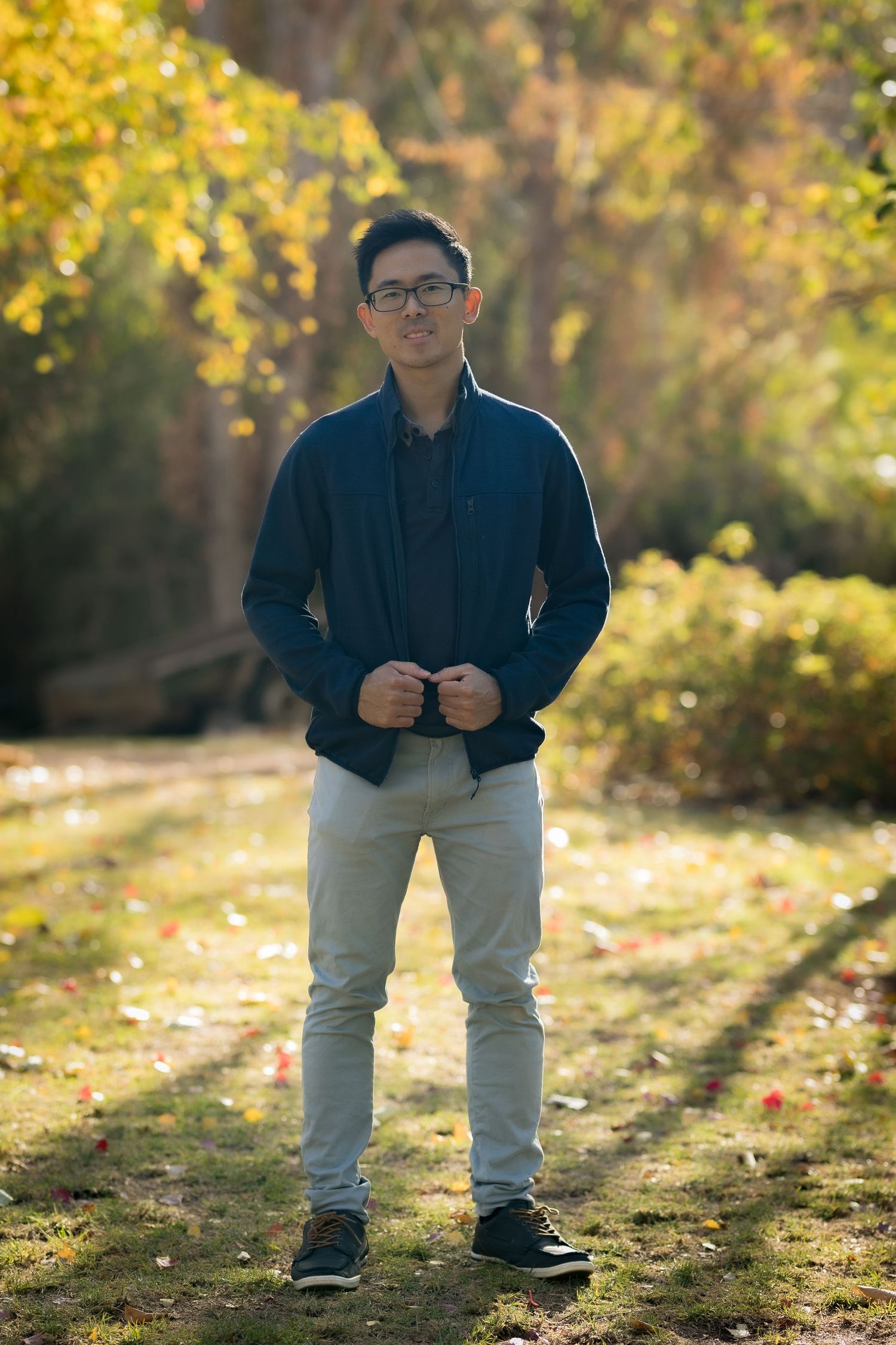 Young Asian man in blue jacket and gray pants standing in a sunlit autumn park with colorful foliage.
