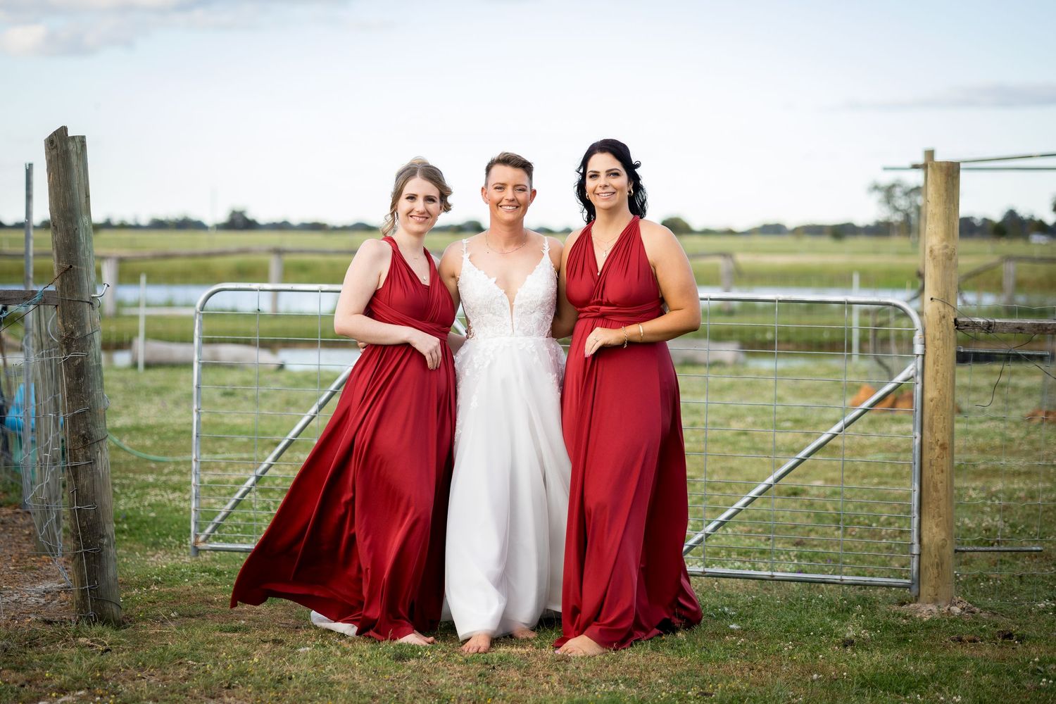 Bridal party in burgundy and white poses casually by rustic fence in countryside setting.