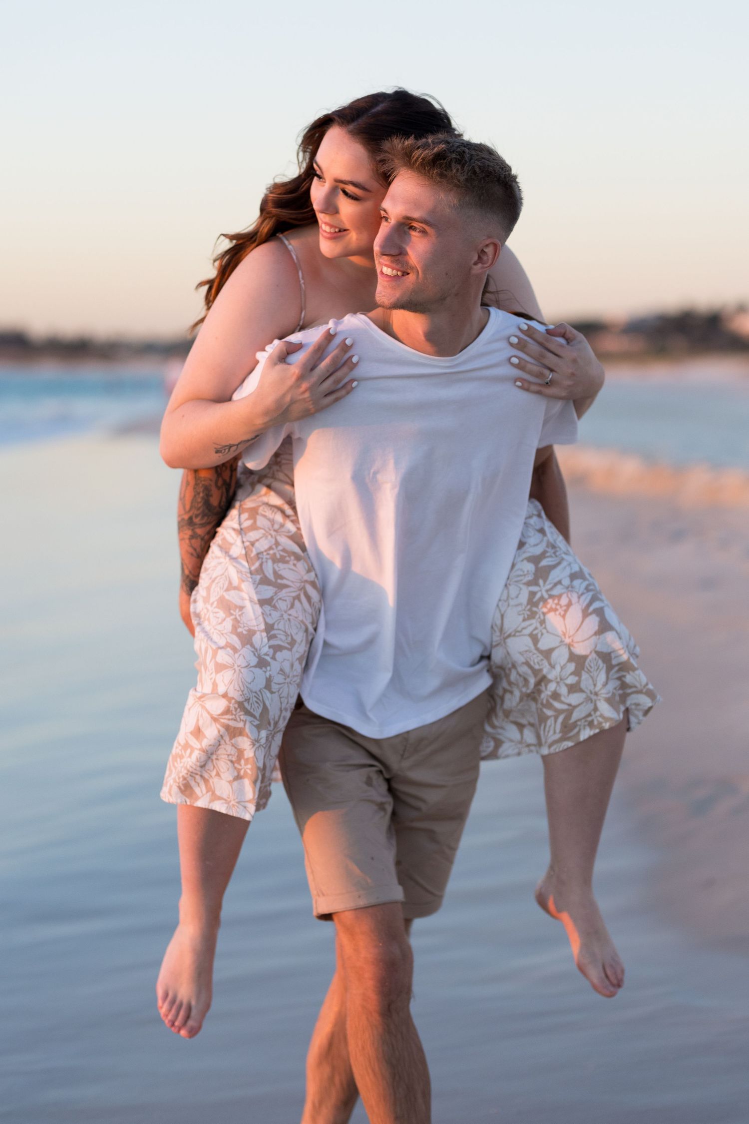 A happy couple enjoys a piggyback ride on the beach at sunset, smiling and embracing near the ocean.