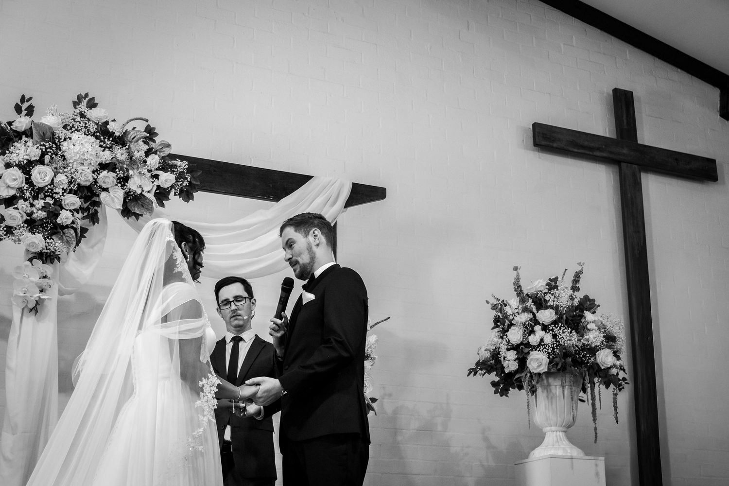 Bride and groom exchange vows at elegant church wedding ceremony with floral arrangements and wooden cross.