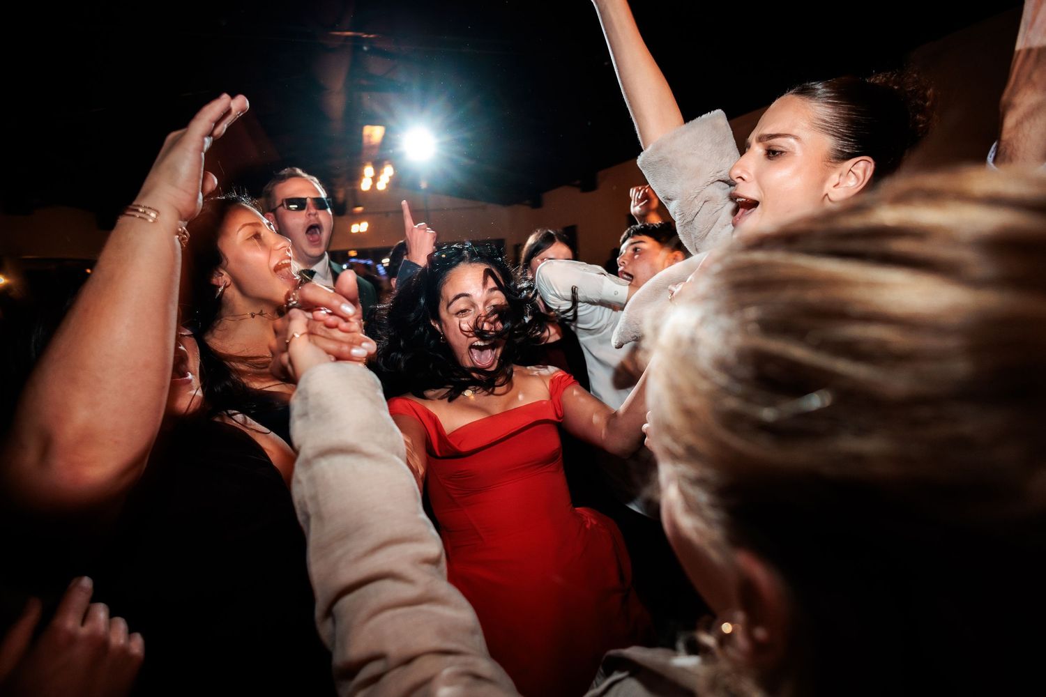 Joyful wedding guests celebrating together at night, with a woman in red dress at center of lively dance floor crowd.