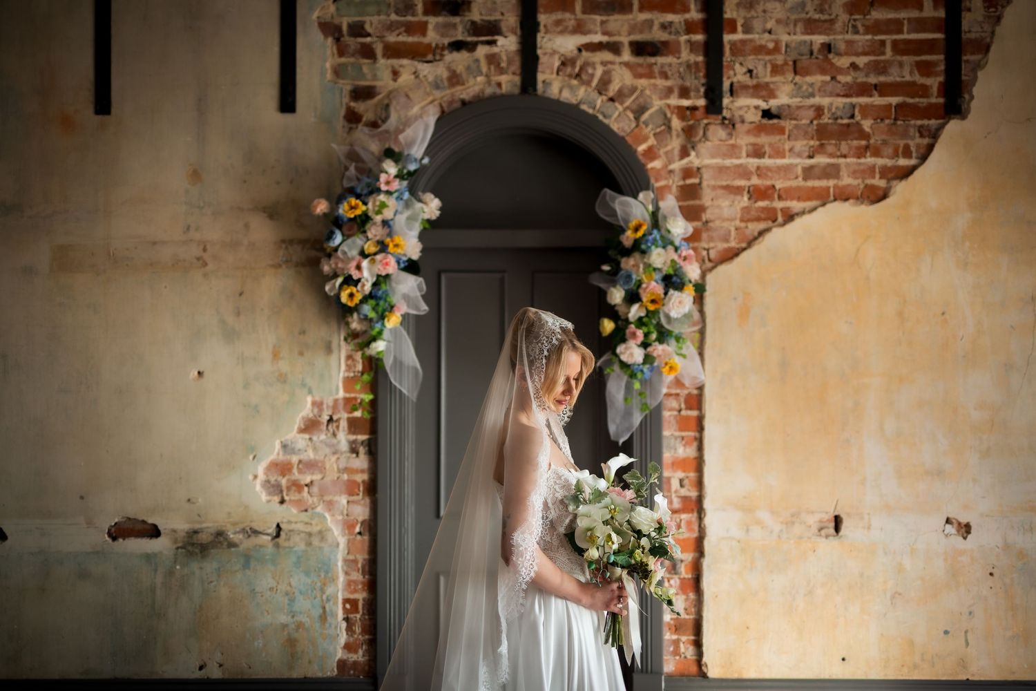 Bride in white gown holding bouquet stands before floral arch on rustic brick wall backdrop.