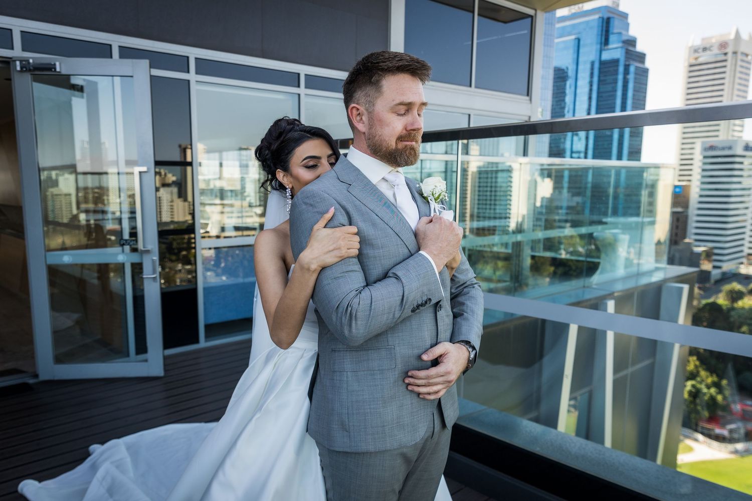 Bride in white gown embraces groom in gray suit on urban balcony with city skyline backdrop.