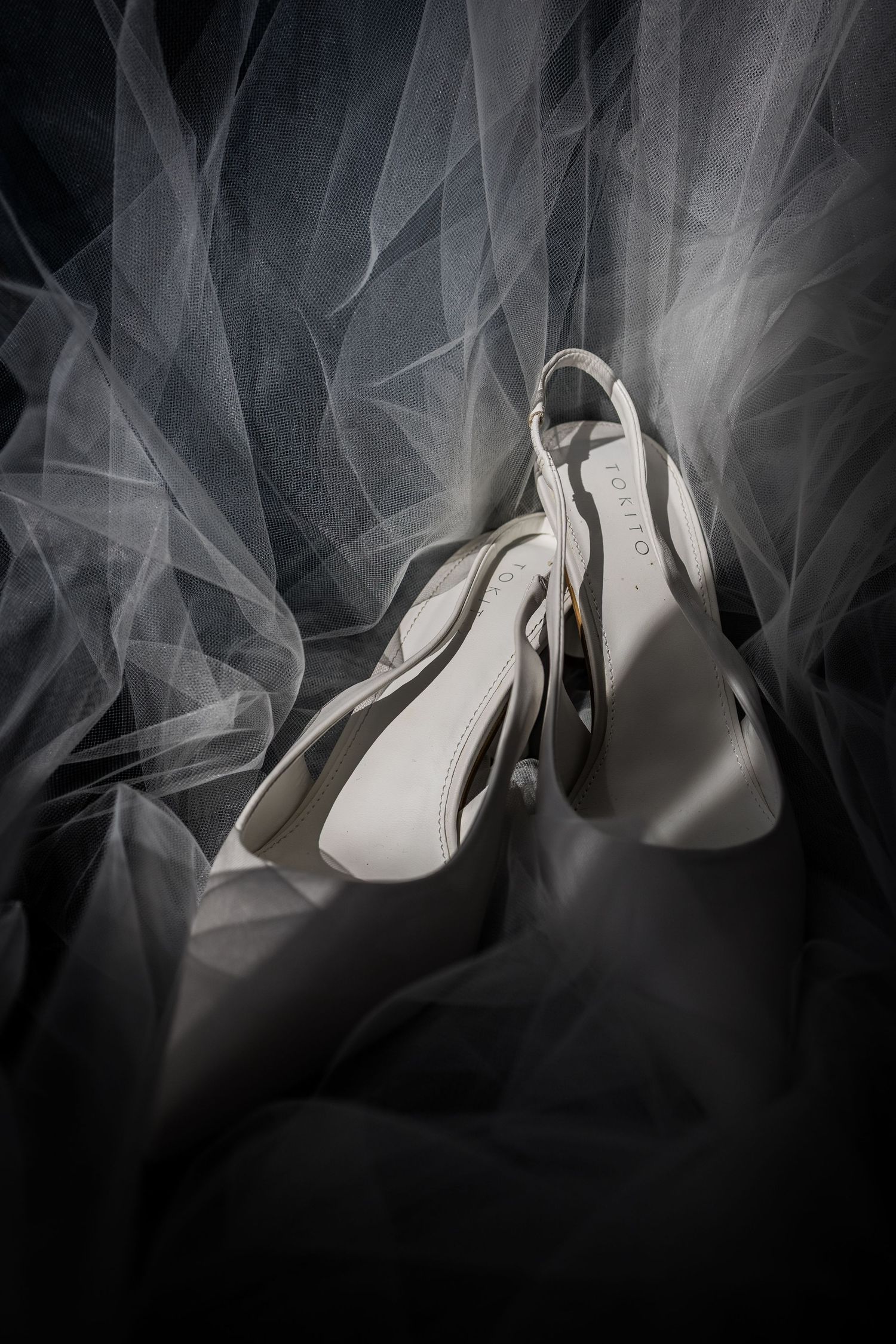 White bridal heels resting on a flowing sheer wedding veil in dramatic black and white lighting.
