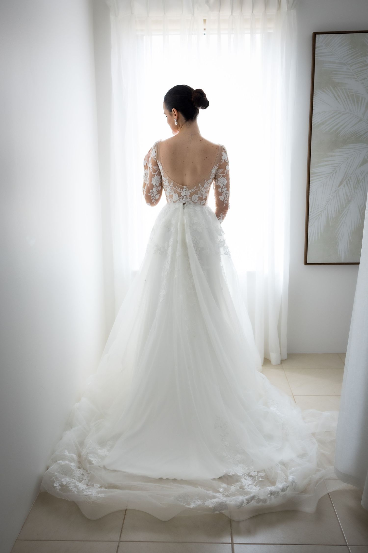 Bride in stunning backless lace wedding gown with floral embroidery stands before a bright window.