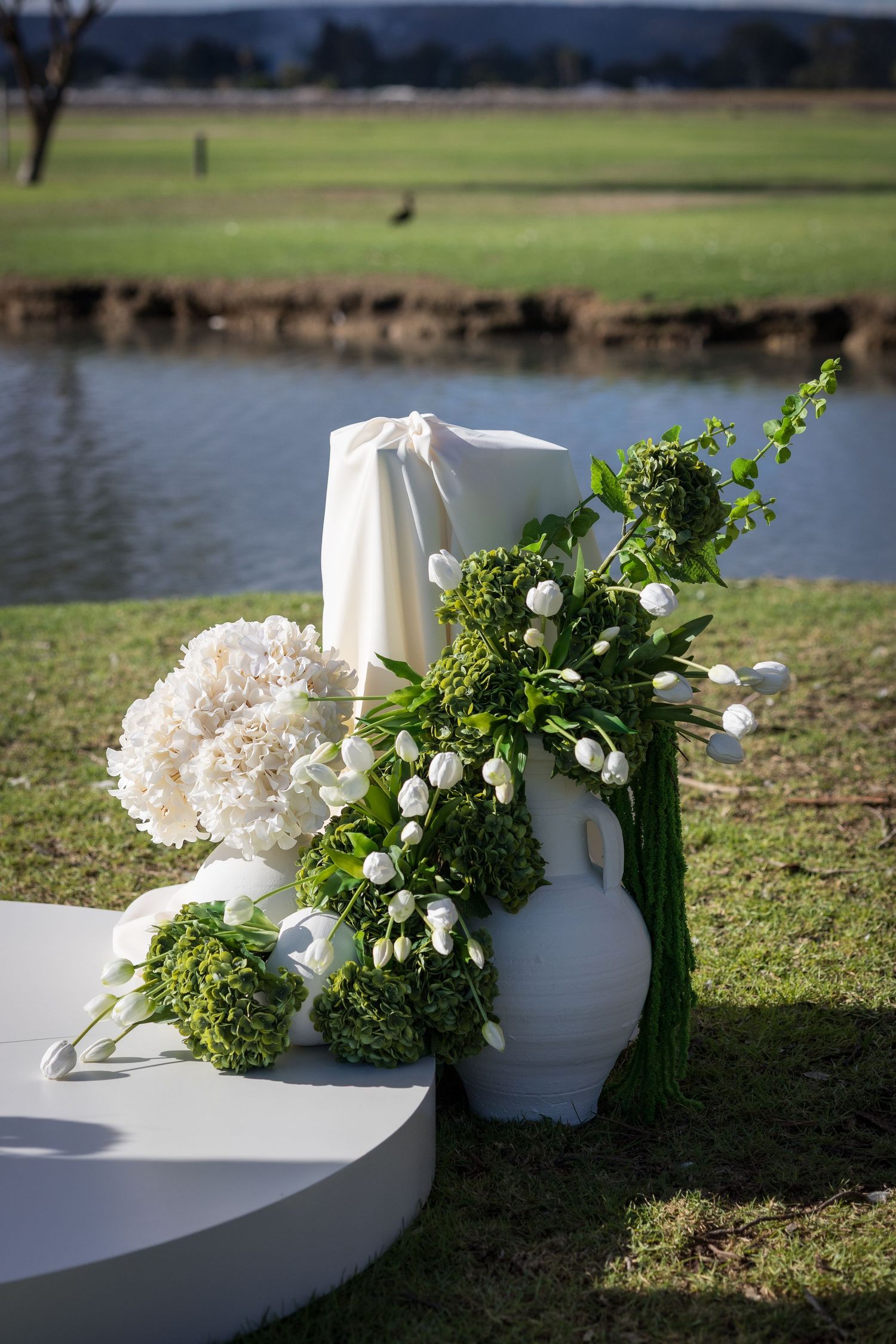 White tulips and hydrangeas in a ceramic vase beside a serene pond at an outdoor wedding ceremony.