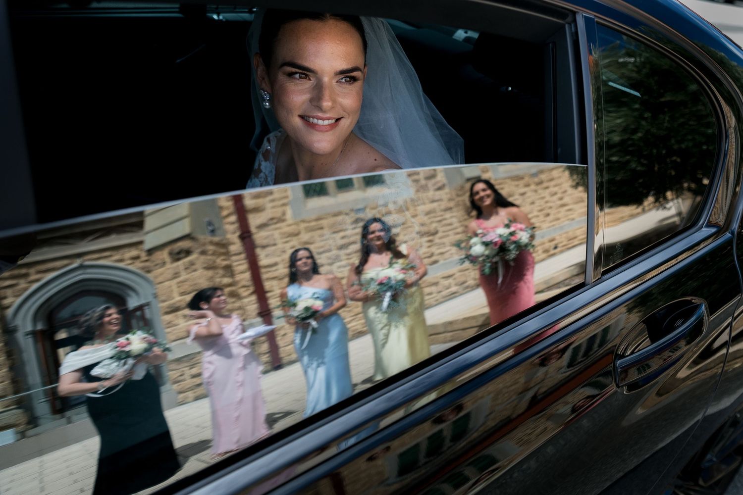 Smiling bride in veil reflected in car window with bridesmaids holding bouquets.