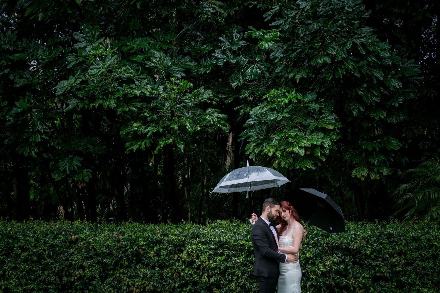 A romantic wedding moment under an umbrella against a lush green garden backdrop.