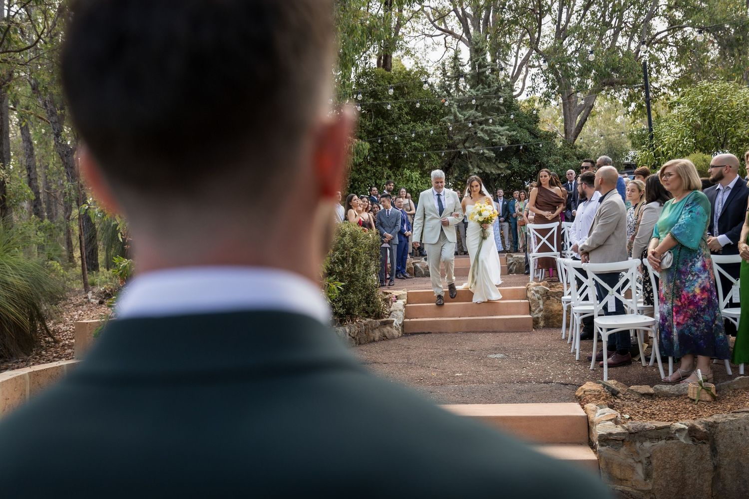 Groom watches bride walk down outdoor aisle surrounded by wedding guests seated on white chairs.