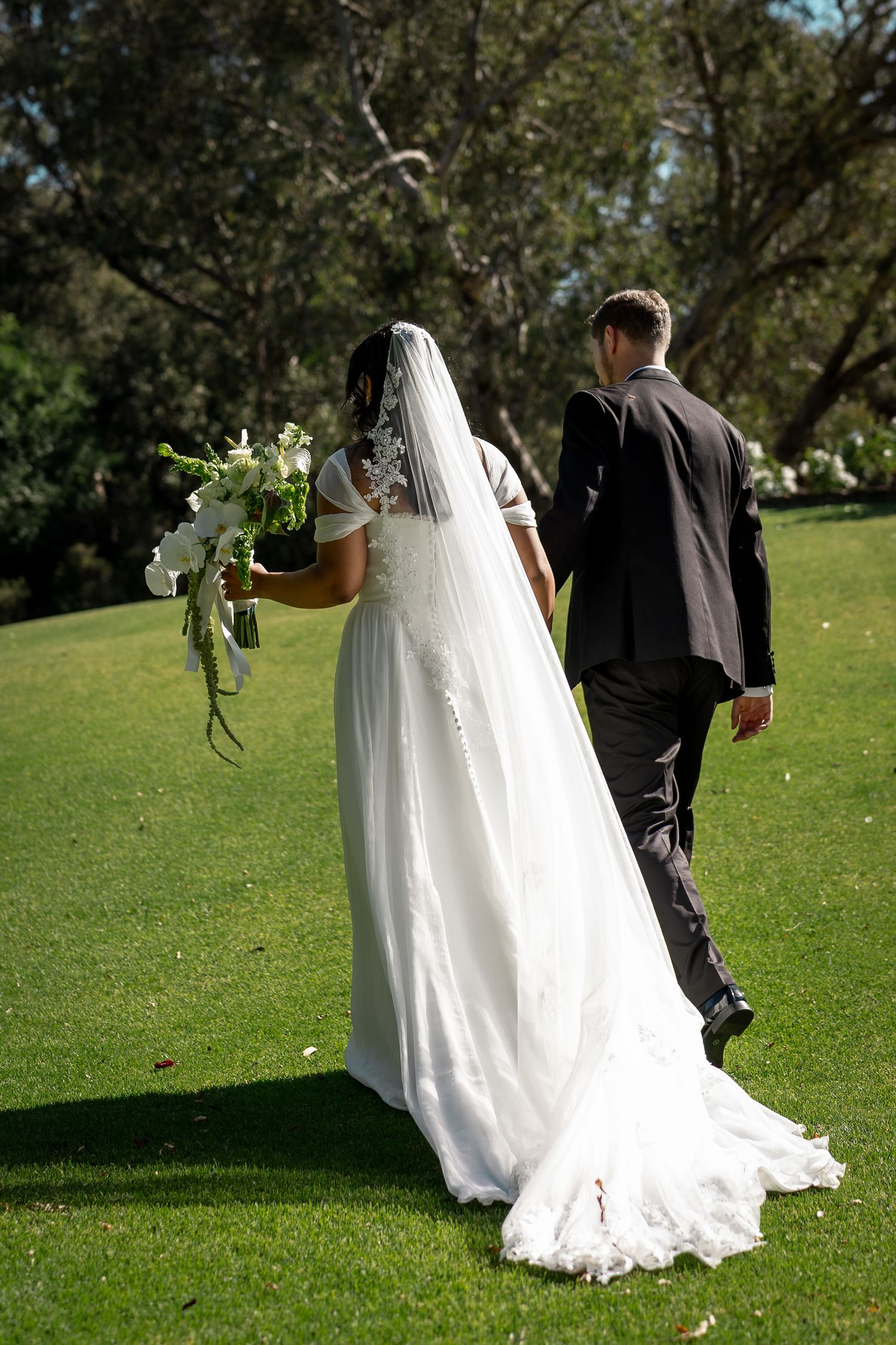 Bride and groom walking hand in hand across green lawn, bride wearing white gown with veil holding floral bouquet.