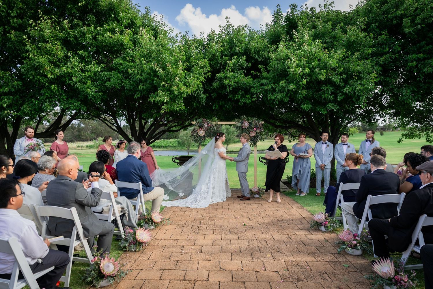 Outdoor wedding ceremony under lush green trees with guests seated on white chairs along a brick pathway.