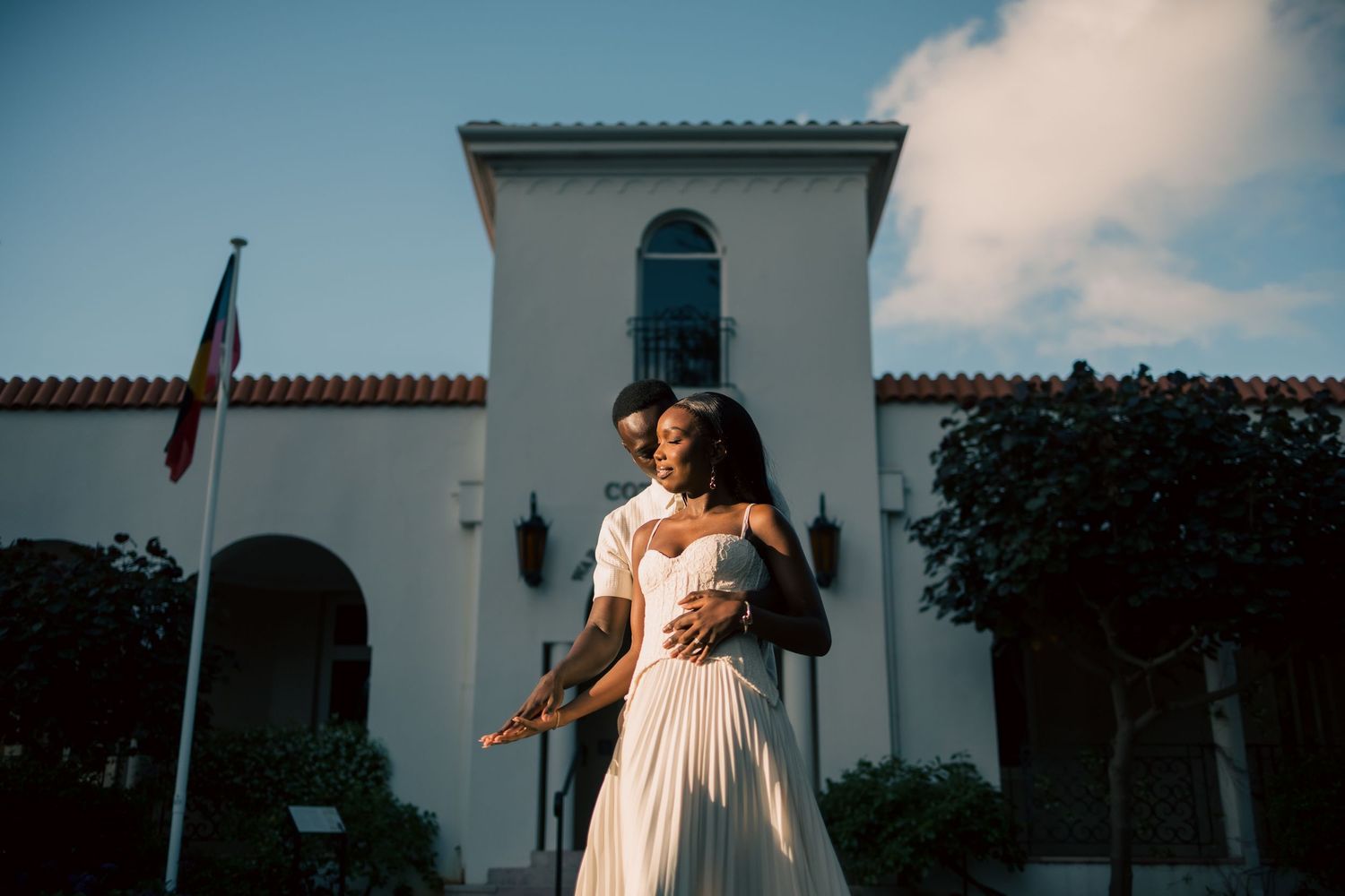 Couple in a romantic embrace at sunset, the woman in a flowing white dress, in front of a white Spanish-style building with a flag.
