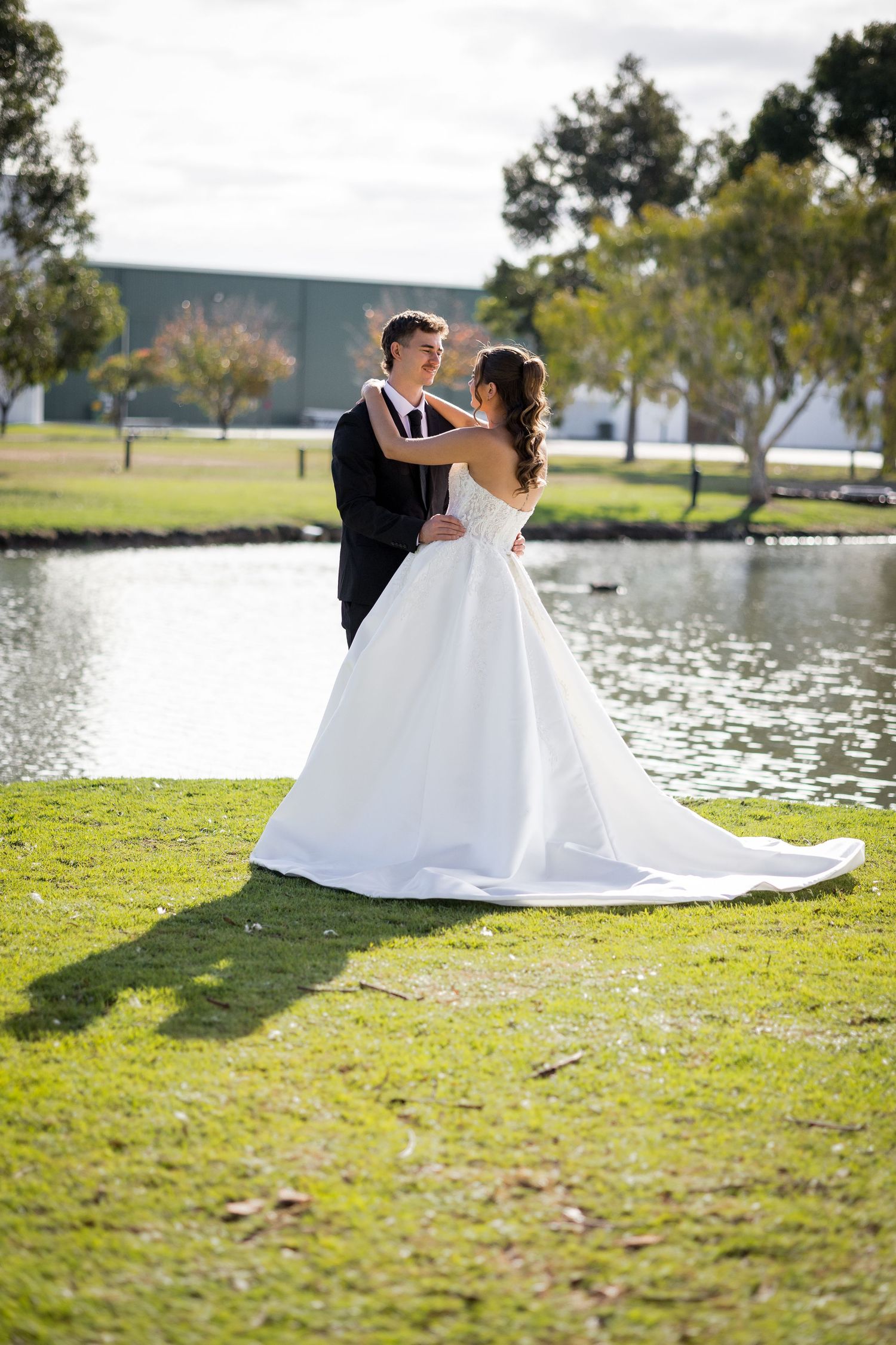 Bride and groom embracing by a serene pond on green grass, bride in elegant white gown with long train.