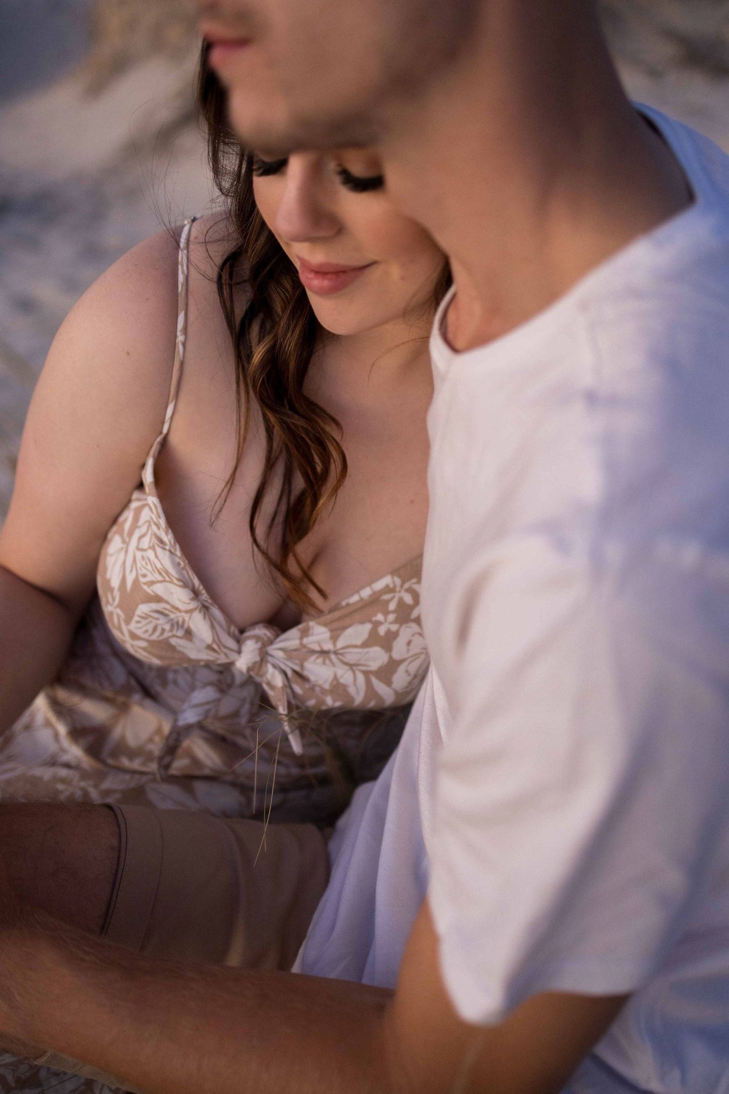 Close-up of couple sitting on sandy beach at golden hour, woman in floral dress smiling softly.