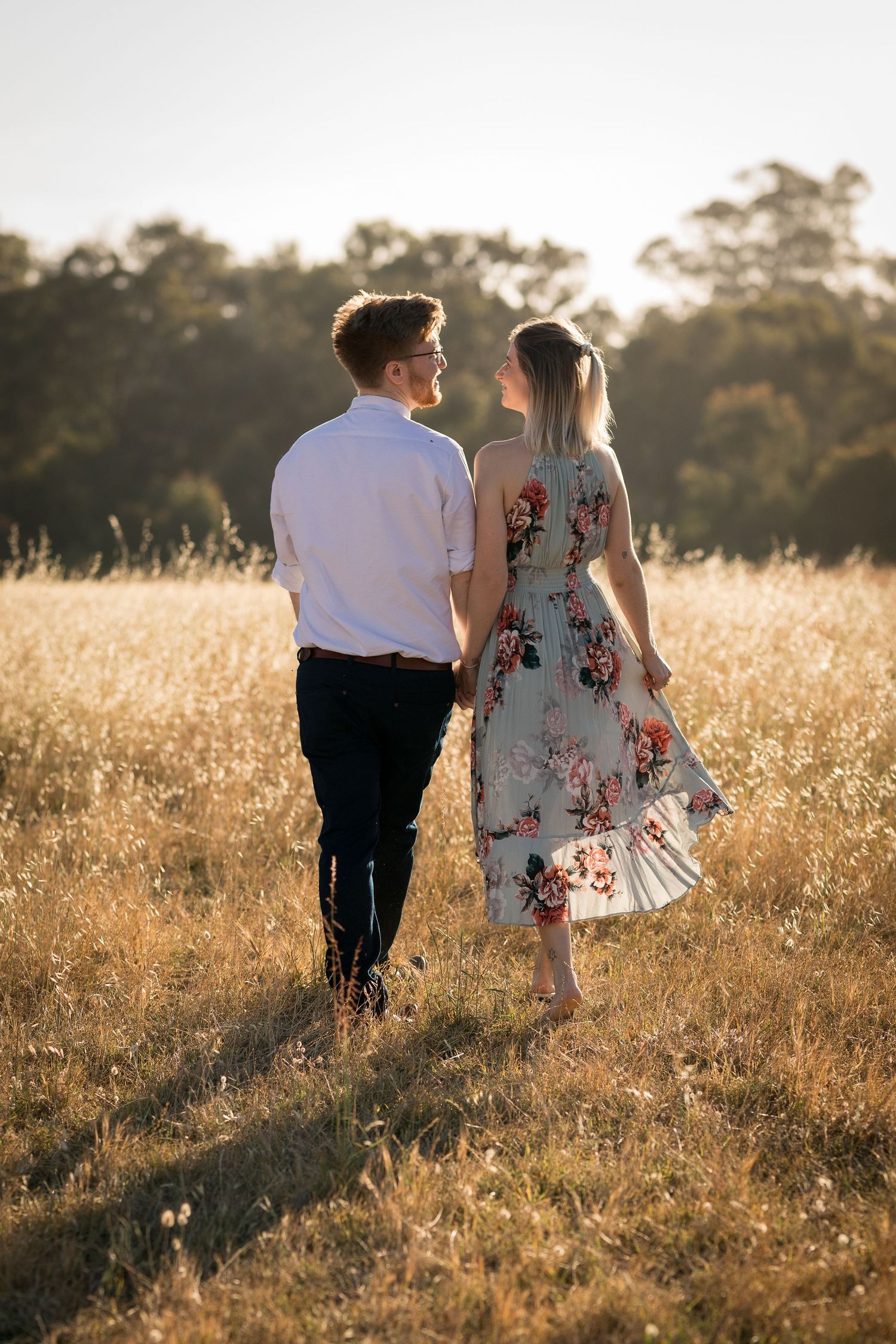A couple strolls hand in hand through tall golden grass during magic hour.