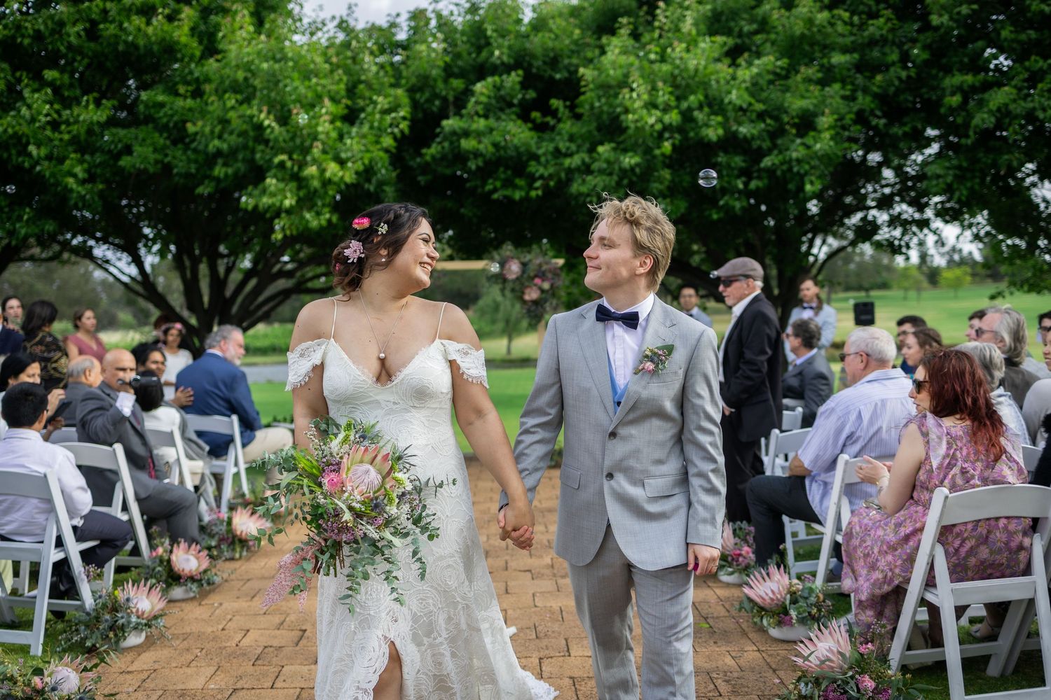 A newlywed couple walks down the aisle at an outdoor garden wedding ceremony surrounded by white chairs and guests.