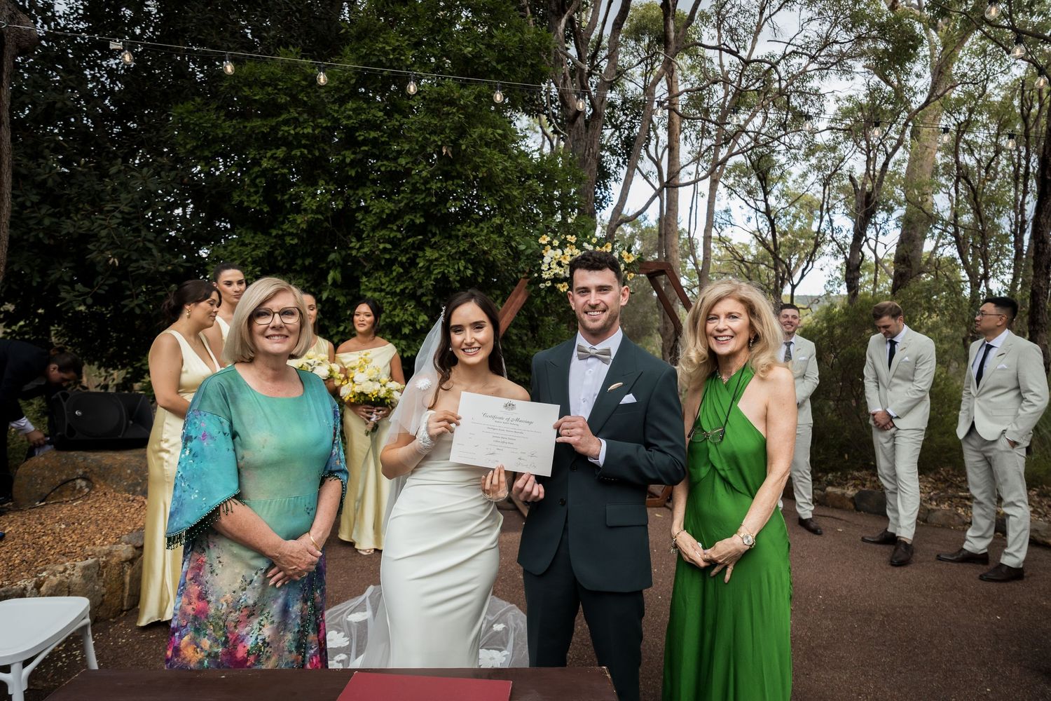 Newlyweds display marriage certificate with officiant and guest at outdoor garden wedding ceremony.