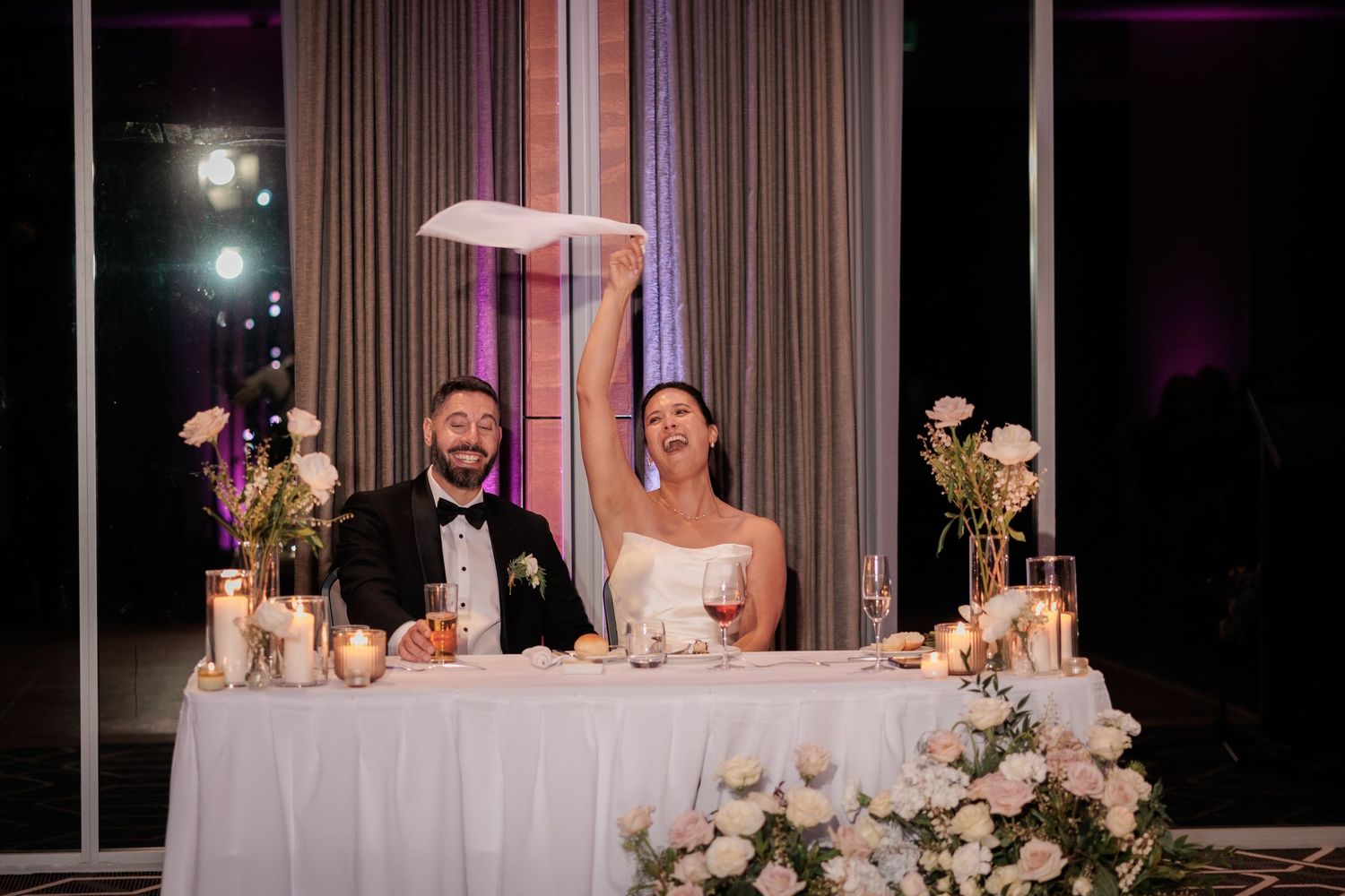 Bride raises napkin in celebration at wedding reception sweetheart table with groom, candles, and white floral arrangements.
